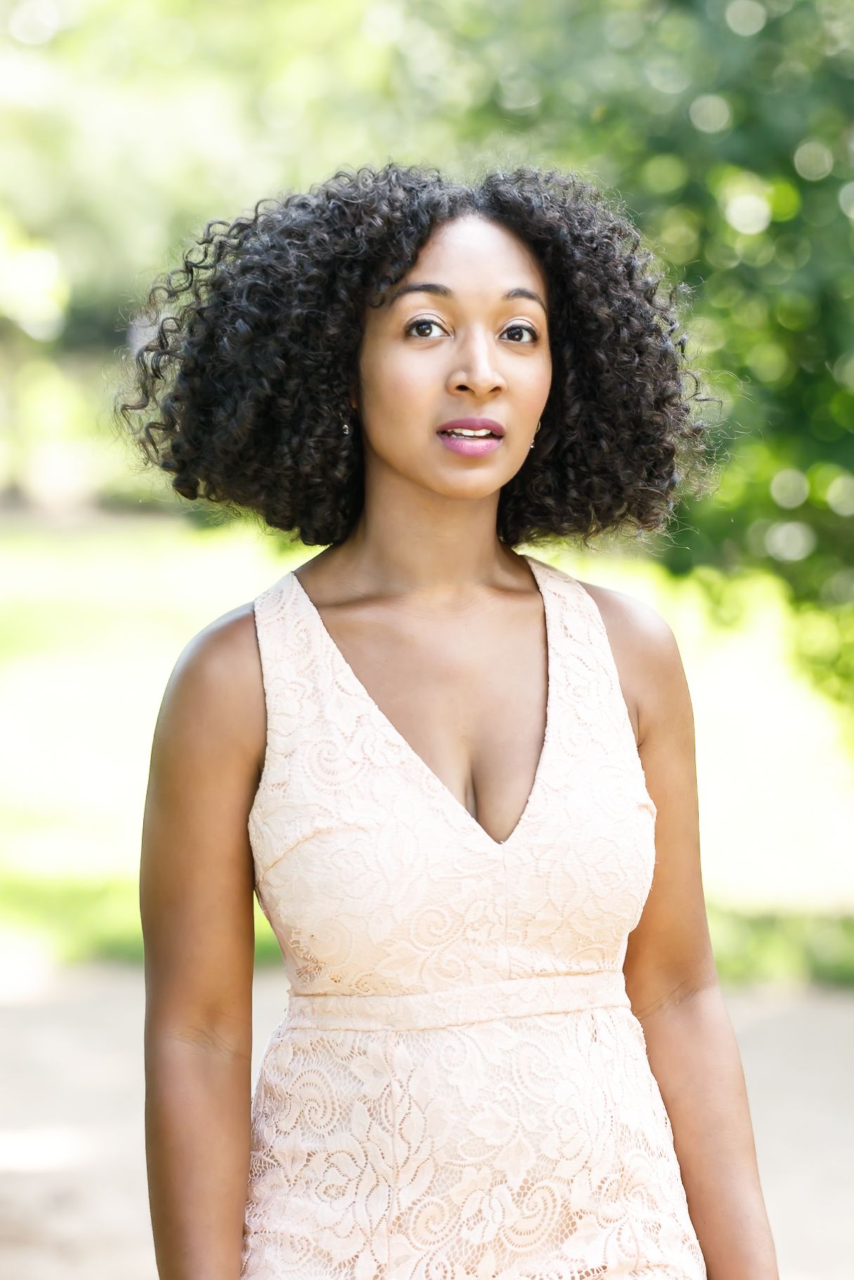 Person with natural curly hair wearing a peach lace dress poses outdoors with soft bokeh background in sunlight.