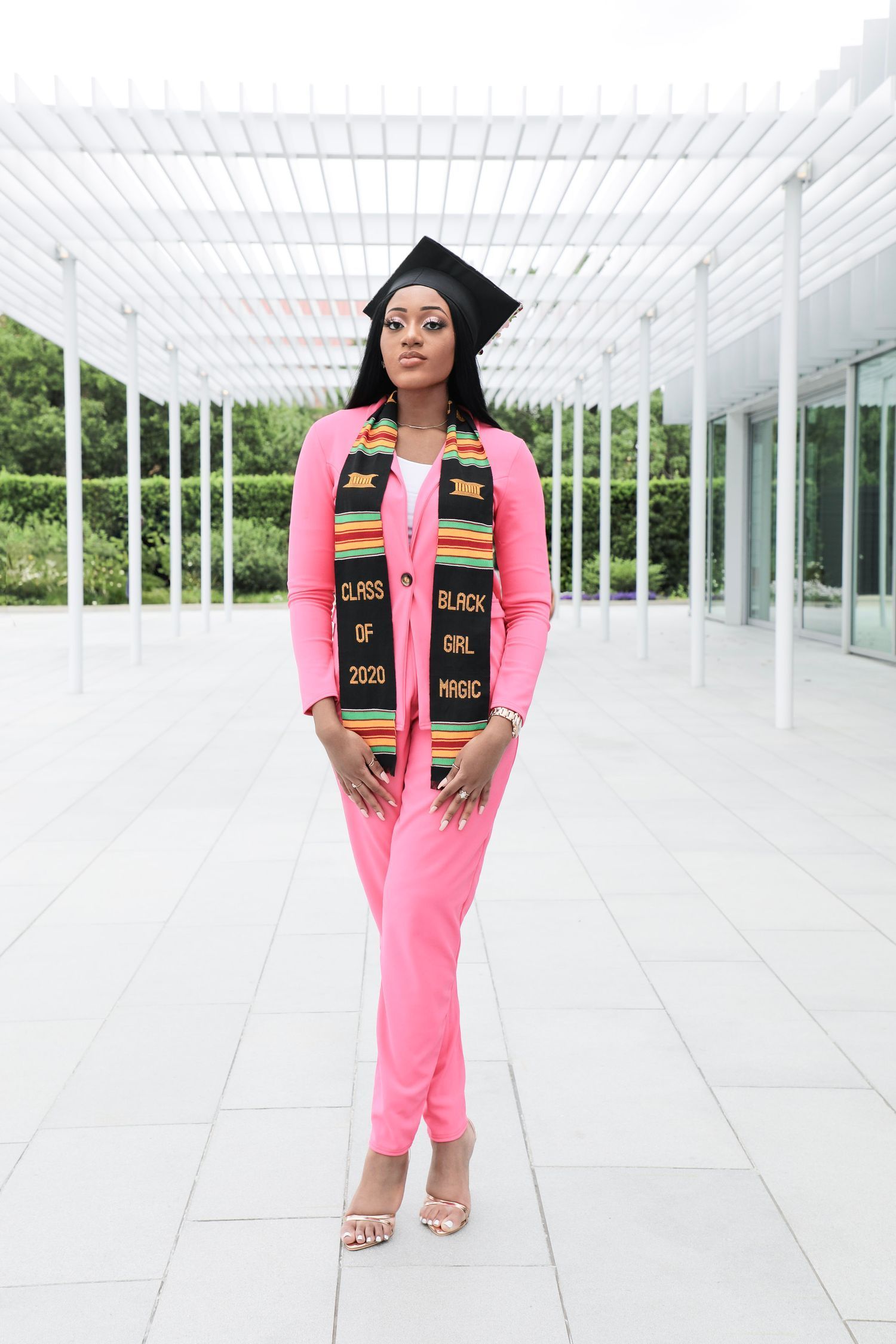 Graduate in pink jumpsuit and graduation cap poses in modern white architectural space with glass ceiling.