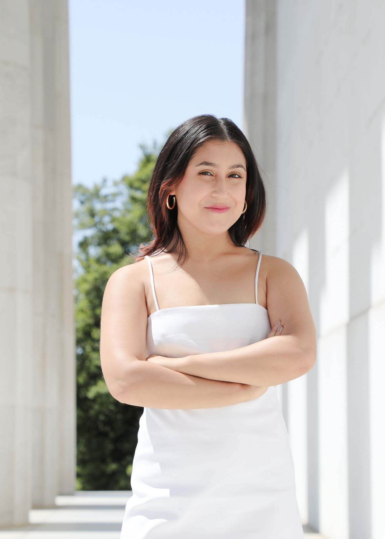 A person in a white spaghetti strap top stands with arms crossed near white columns outdoors on a sunny day.