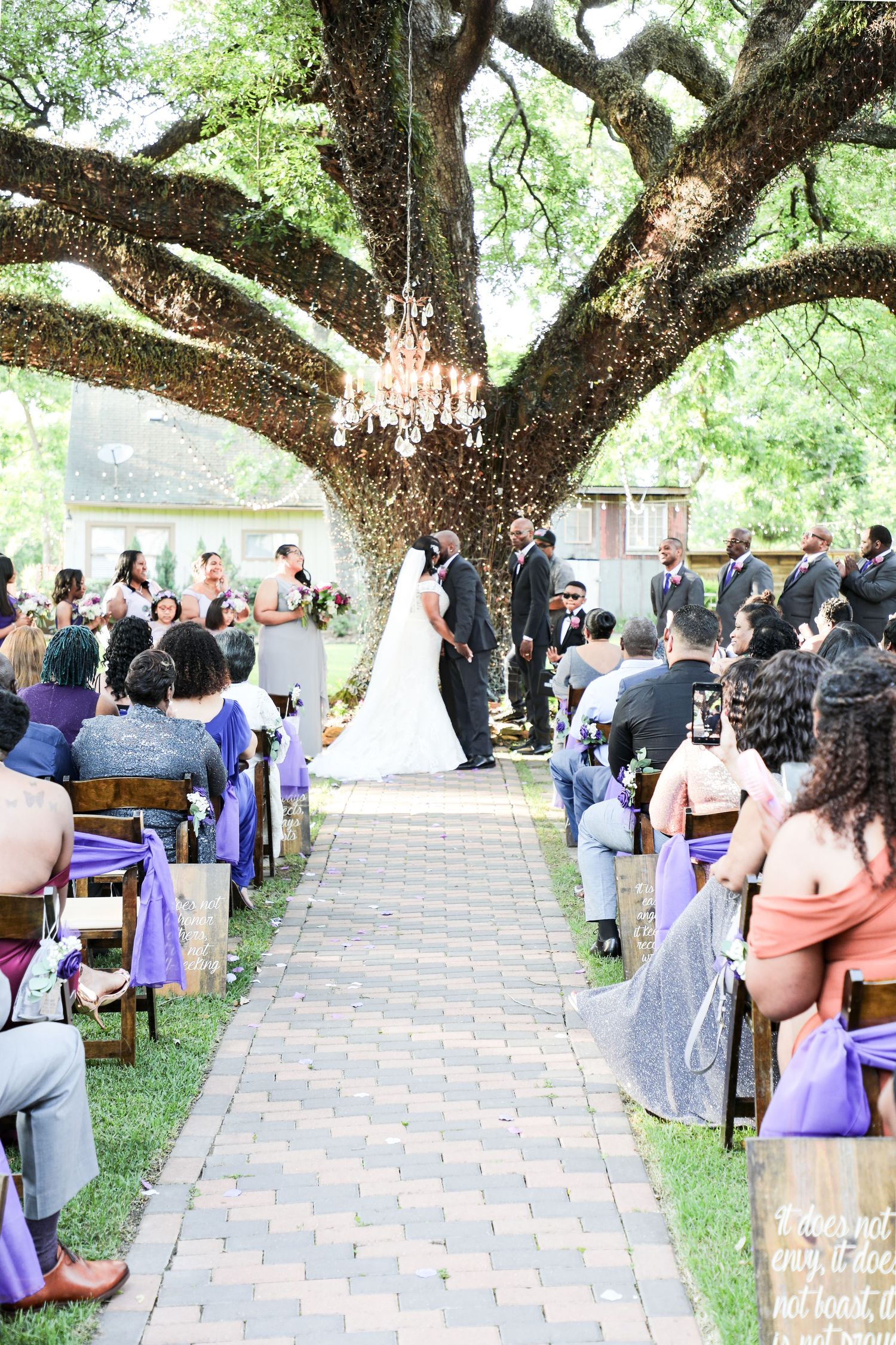 Outdoor wedding ceremony under a large oak tree with chandelier, purple chair sashes, and guests lining a brick aisle.