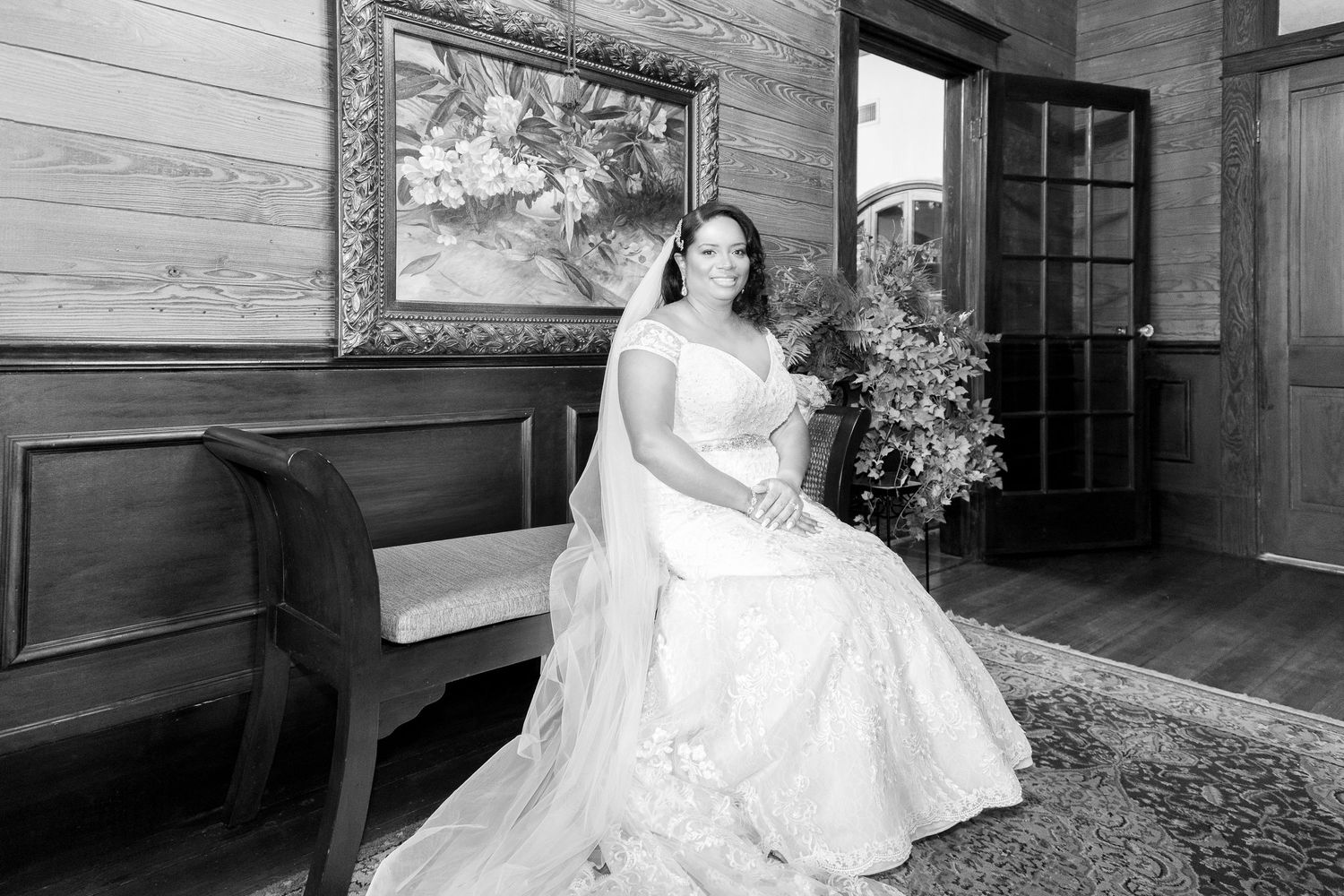 Black and white photo of a bride in a white gown sitting gracefully on a bench inside a rustic wooden venue.