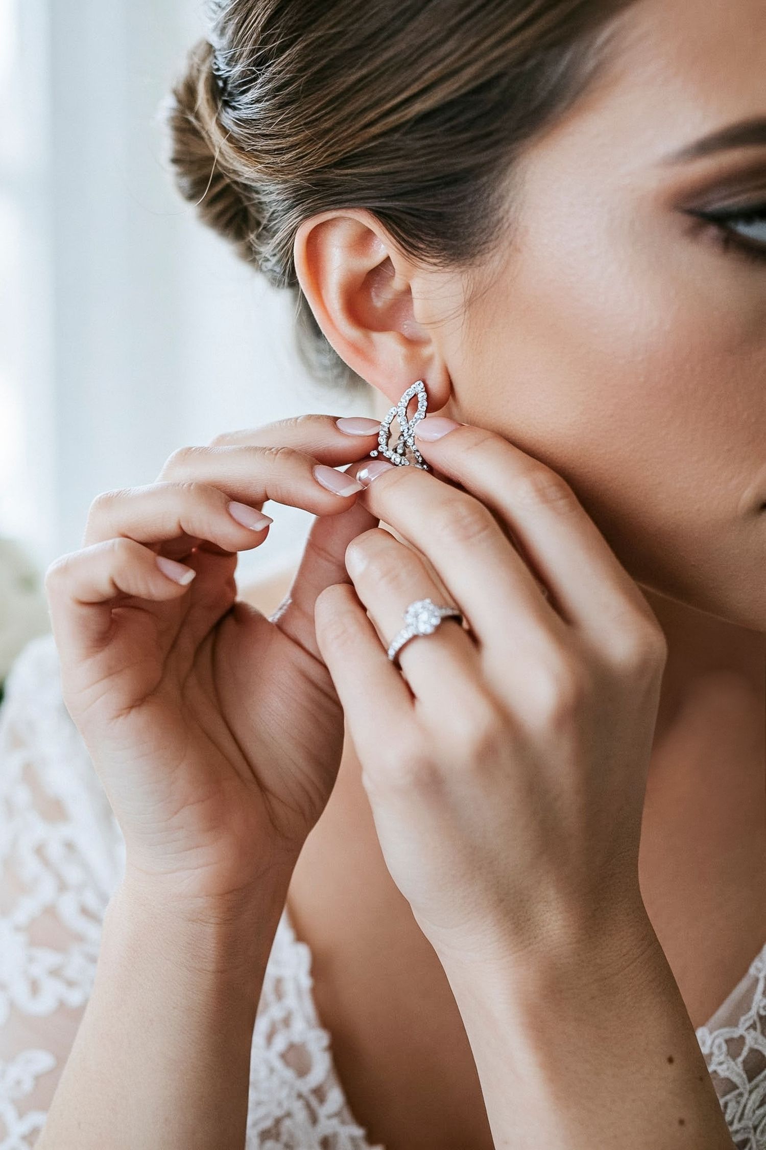 Close-up view of hands putting on delicate earring while wearing lace and beautiful diamond engagement ring.