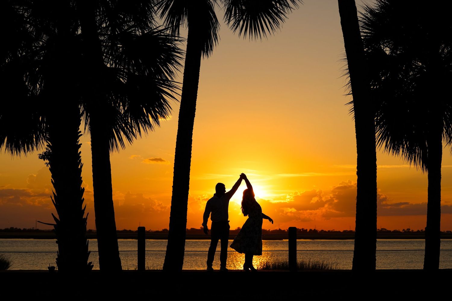 Silhouettes of palm trees and people against vibrant orange sunset sky over tropical beach.
