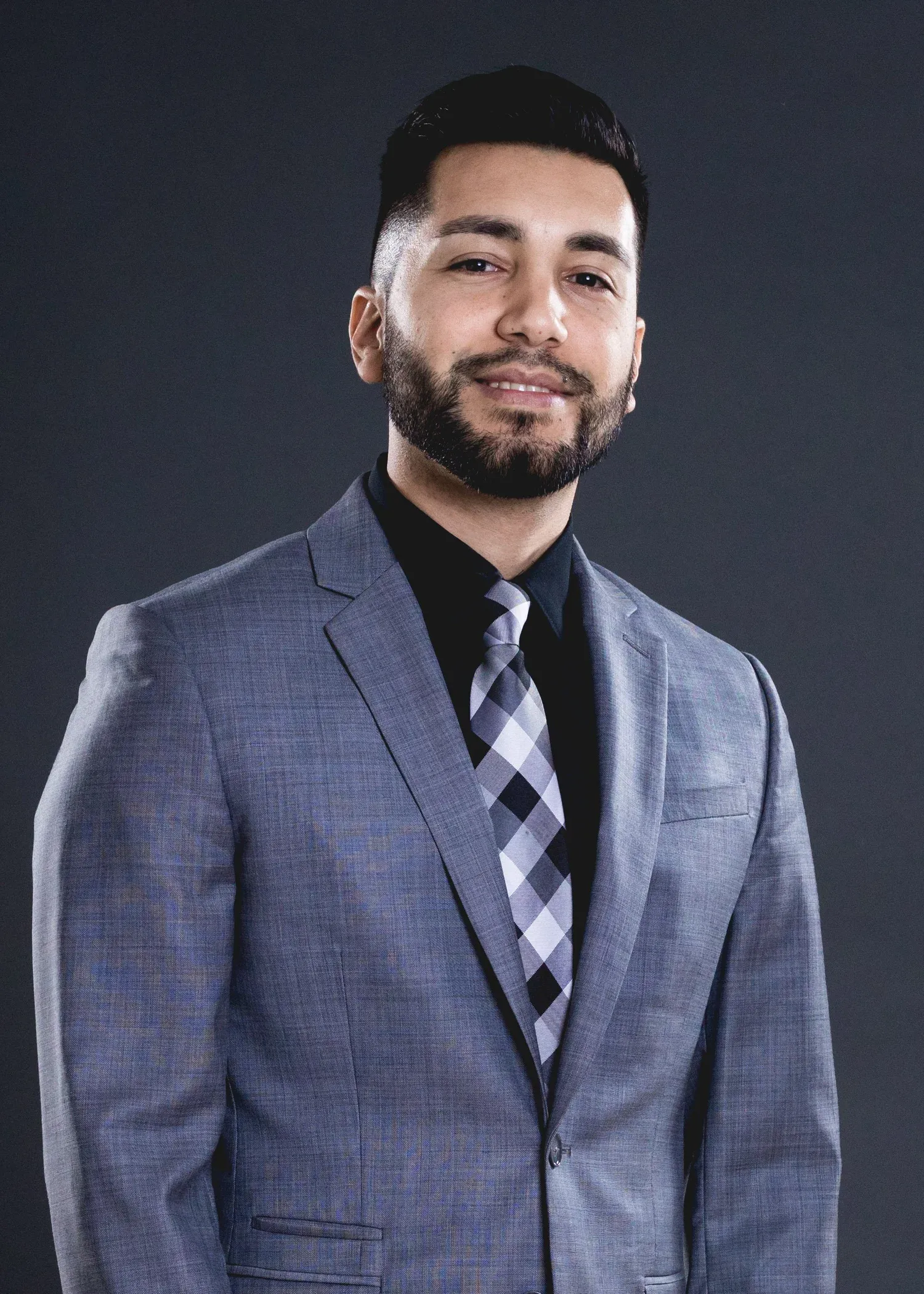 Professional headshot of a wedding videographer wearing a gray suit jacket with black shirt and plaid tie against dark background.
