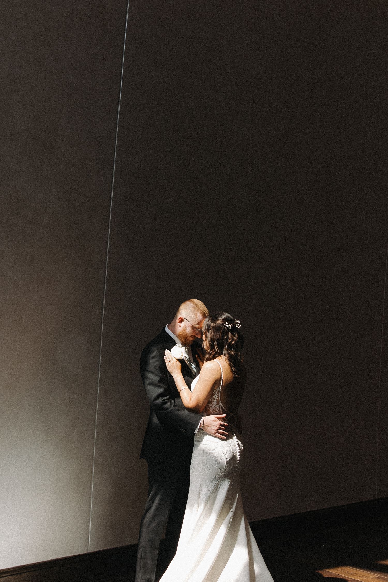 A romantic couple shares an intimate moment in formal wedding attire against a dark minimalist backdrop.
