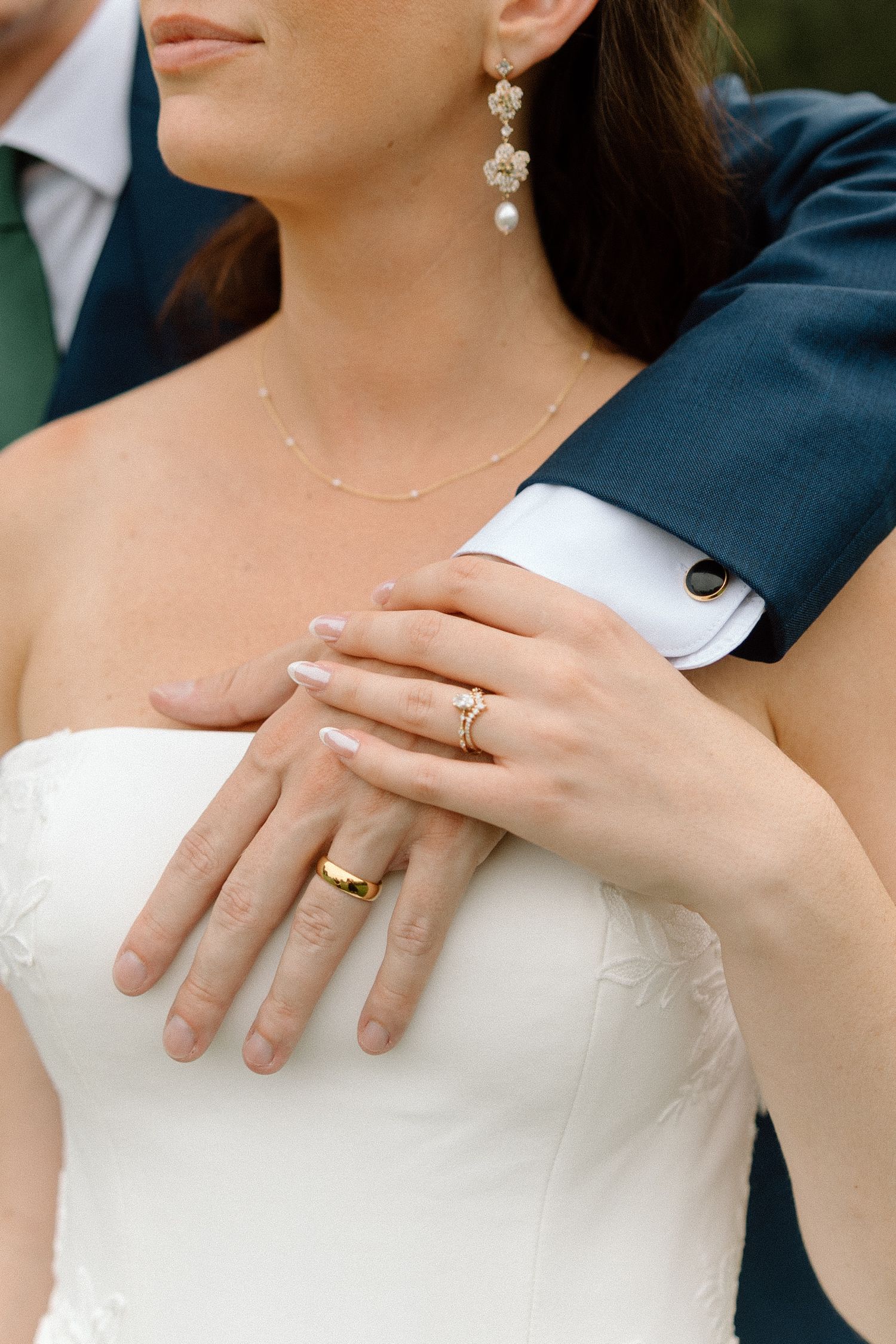 Close-up of wedding jewelry and rings with hands embracing against a white dress and navy suit.