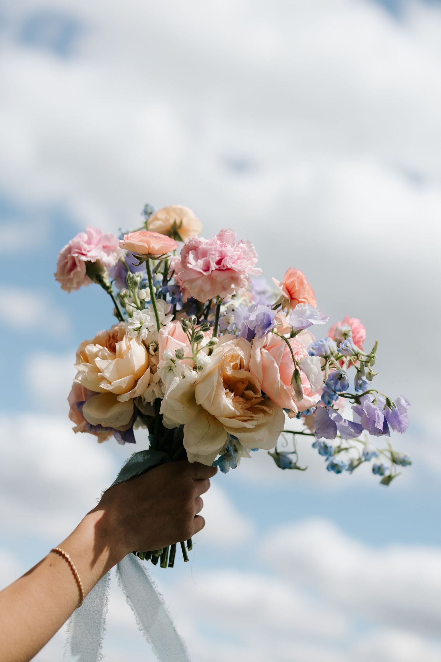Hand holding a bridal bouquet of pink, peach, and blue flowers against a cloudy sky.