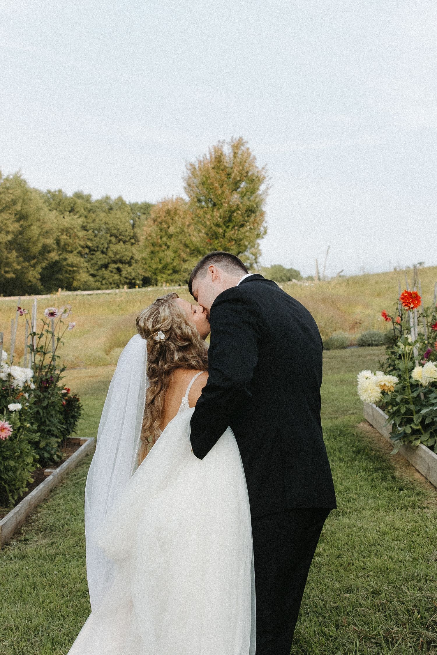 A couple shares their first kiss as newlyweds in an outdoor garden setting with flowers and greenery.