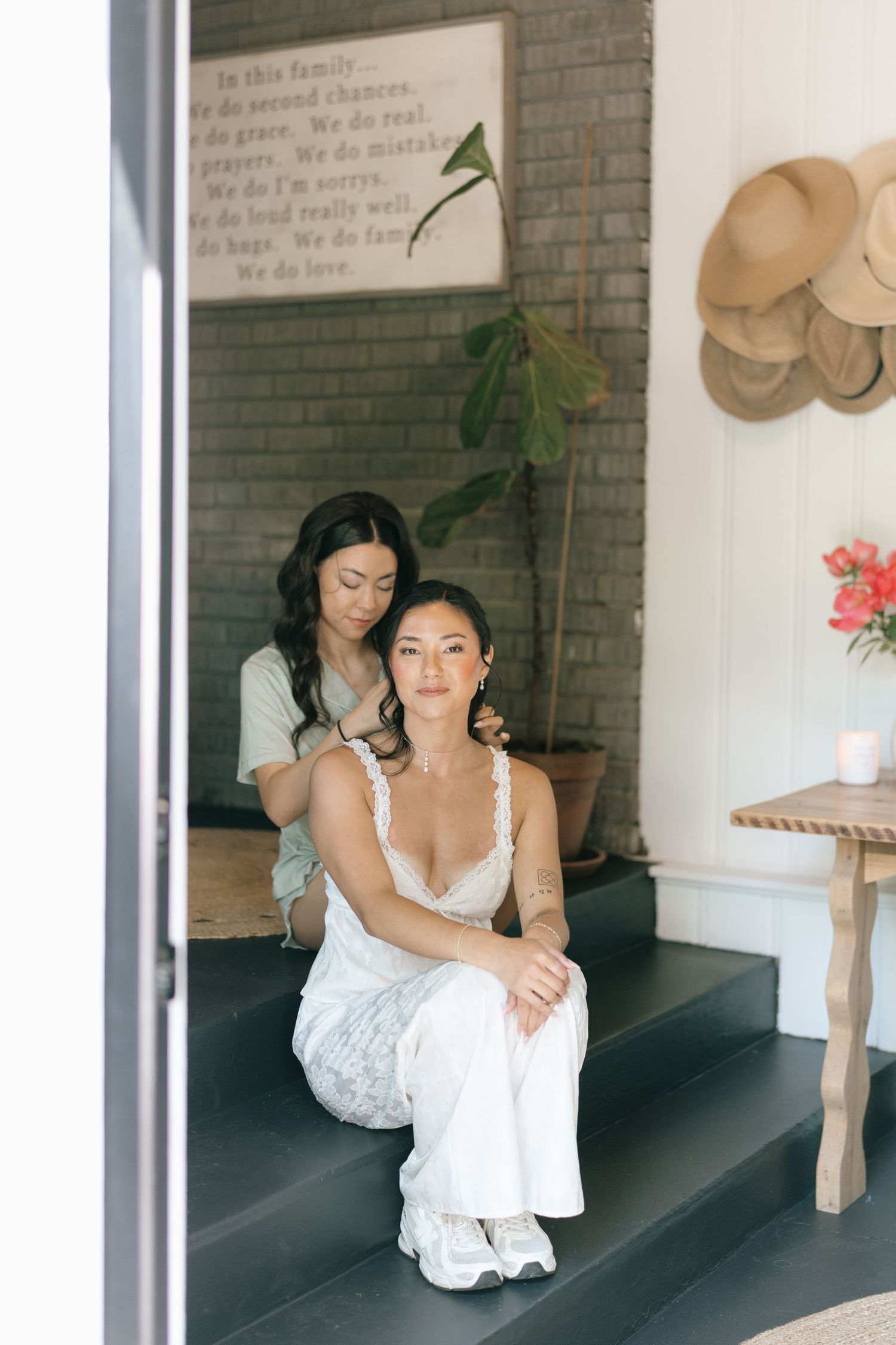 Two women in white outfits sit on dark steps inside a cozy home with hats on wall and motivational sign.