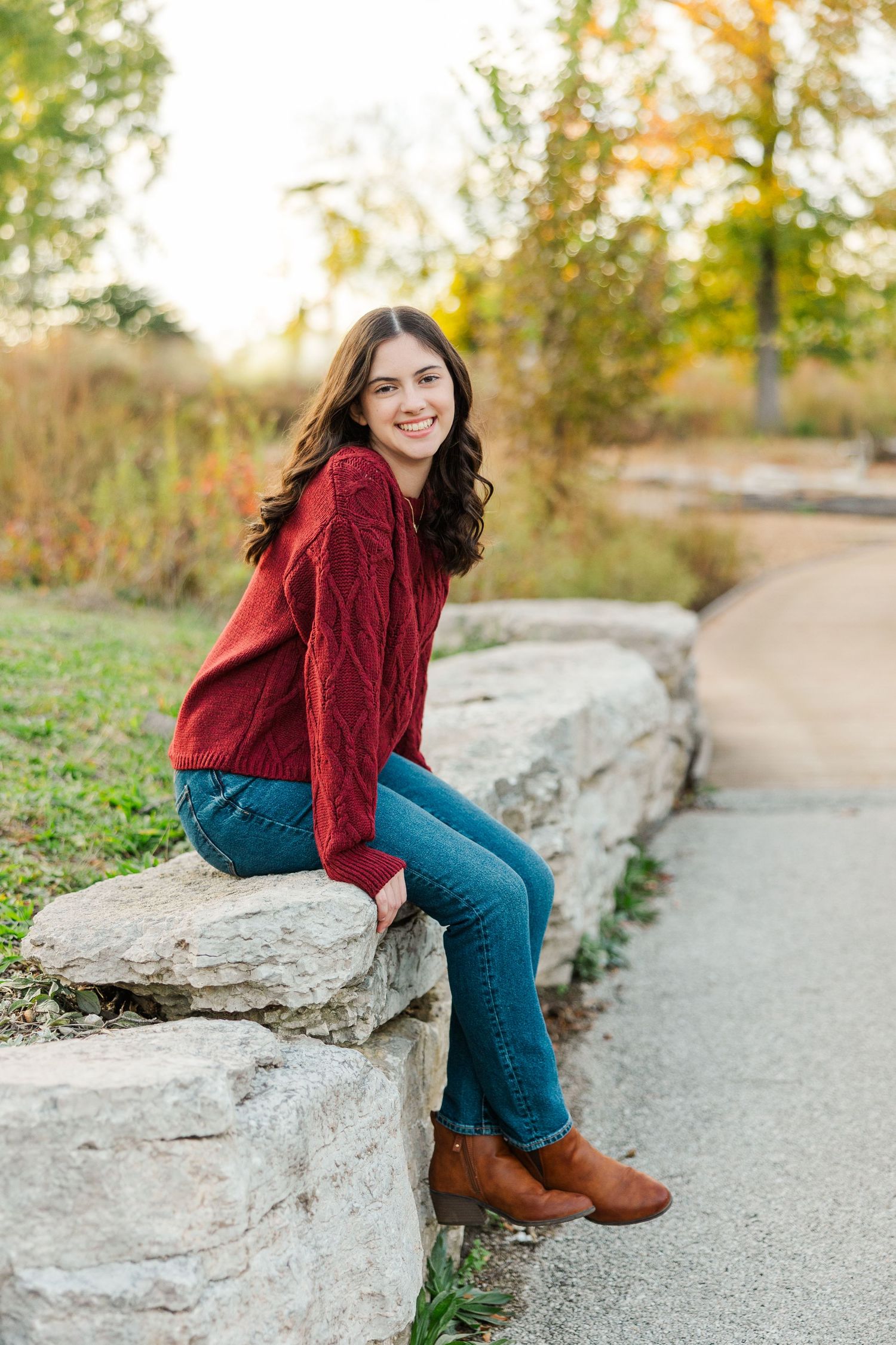 Young woman in red sweater and jeans sitting on stone wall outdoors in autumn park setting.