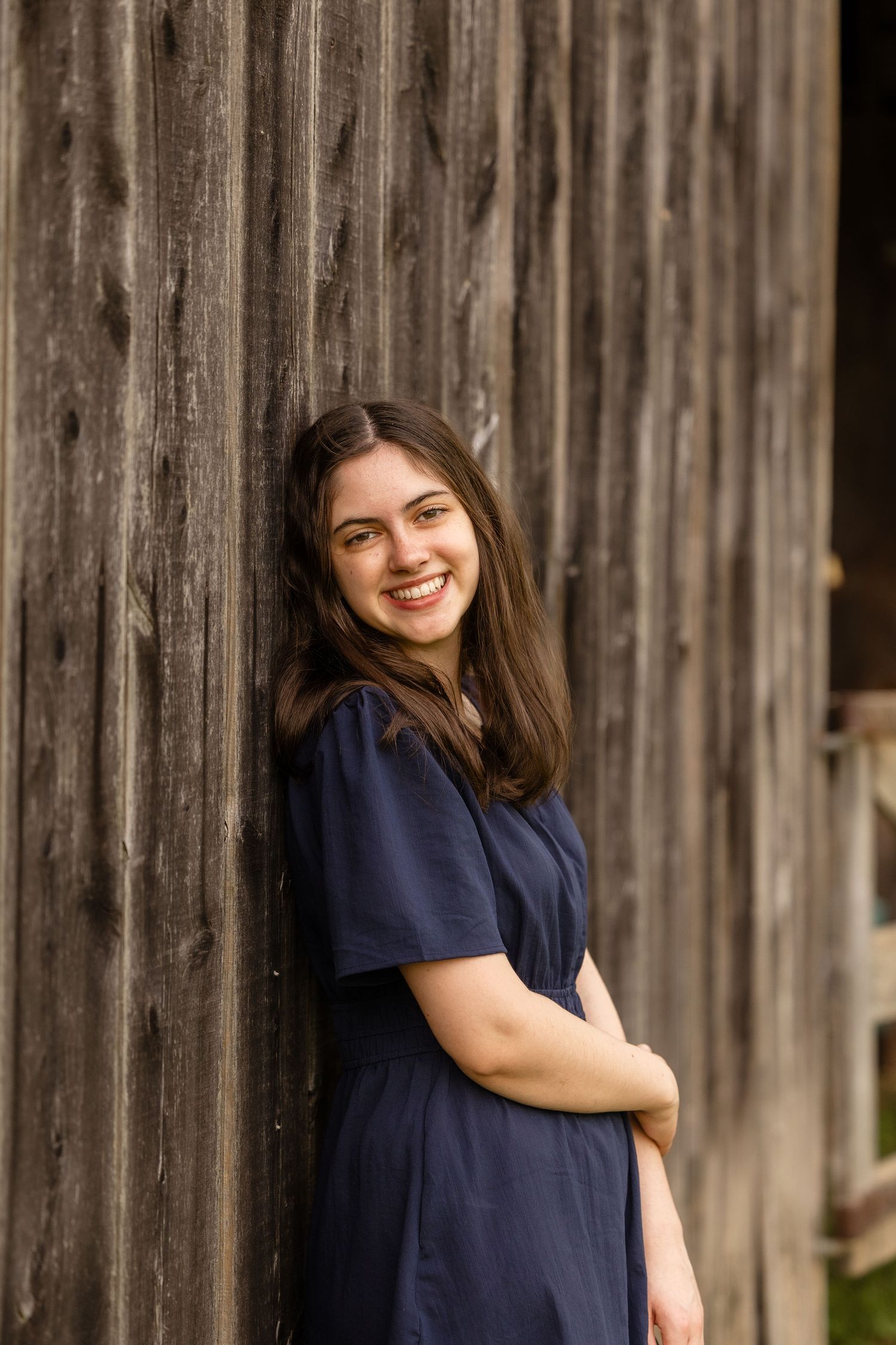 Portrait of someone in a navy dress leaning against rustic wooden wall outdoors.