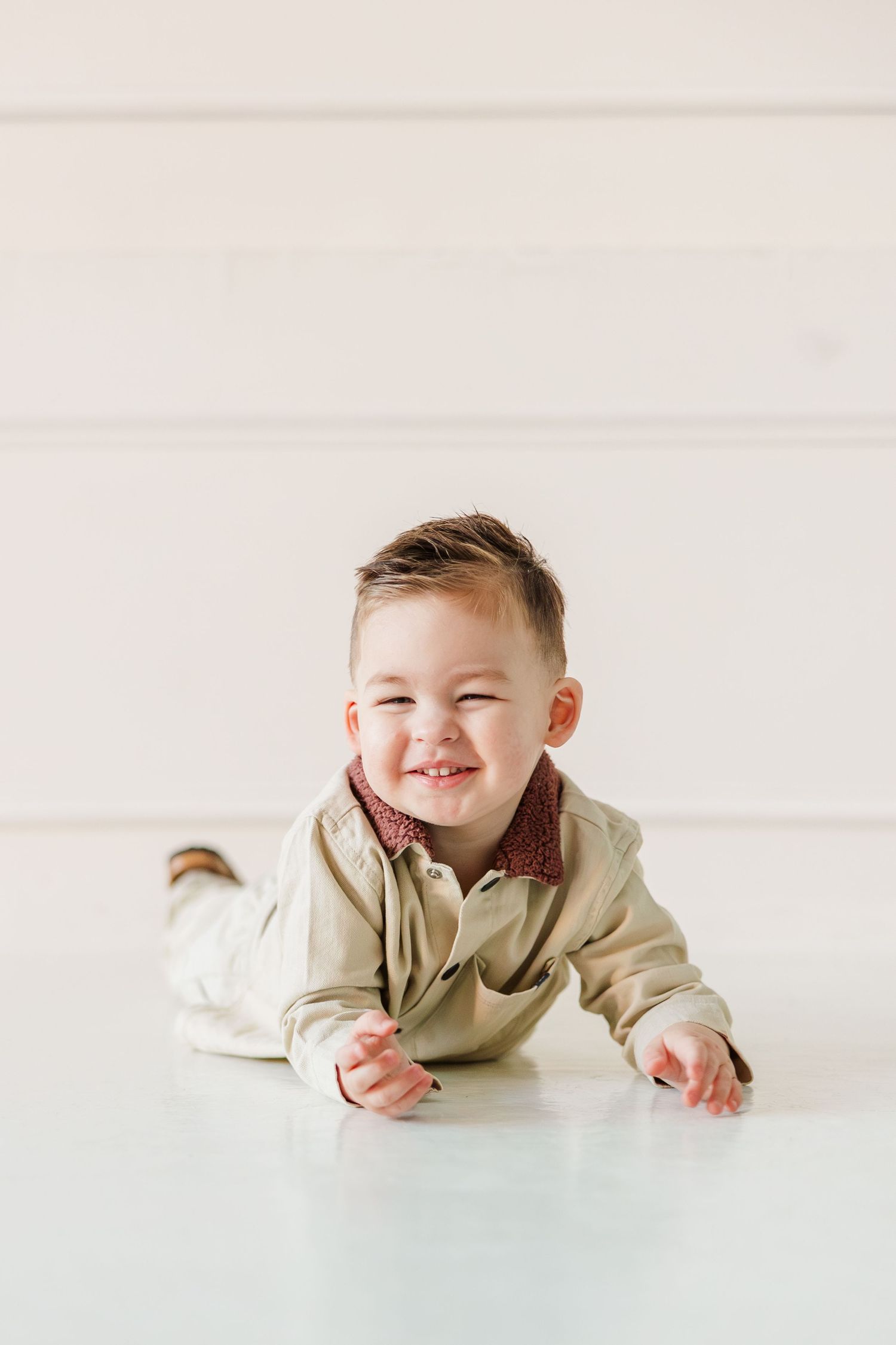 Smiling baby boy in beige outfit lying on white floor, looking at camera with a joyful expression.
