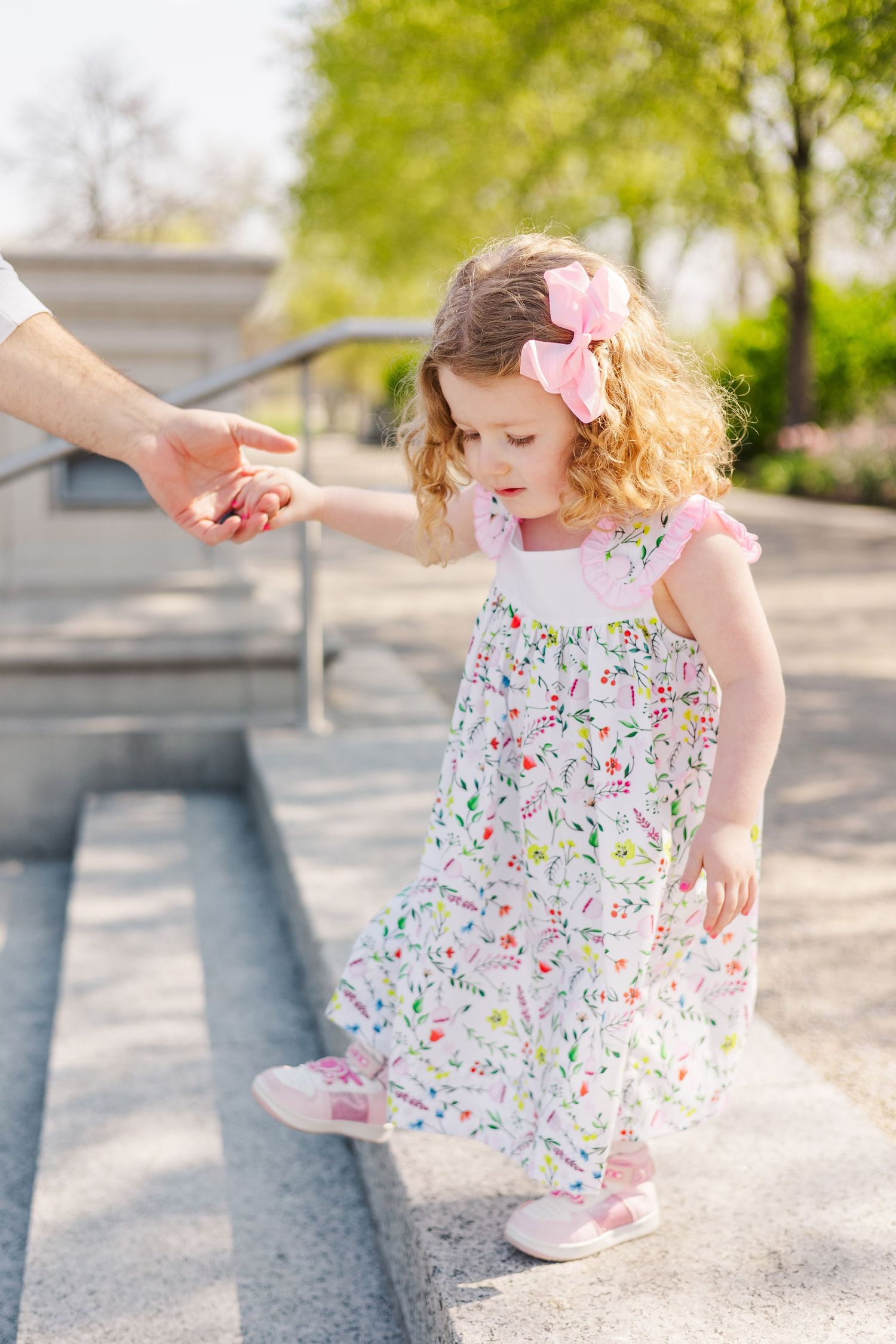 Toddler girl in floral dress with pink bow holds adult hand descending outdoor steps on sunny day.