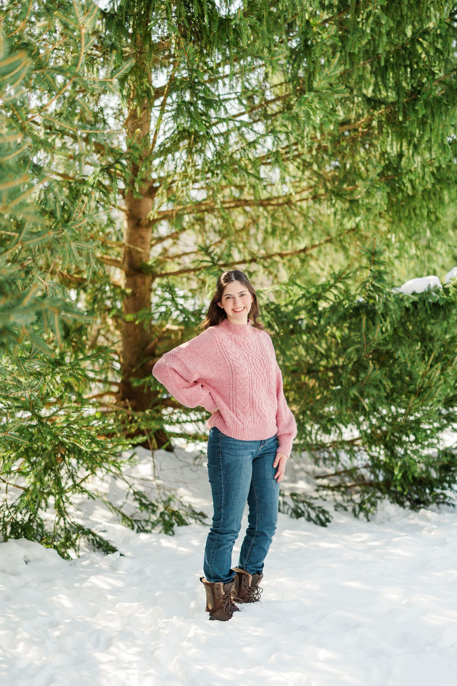 Person in pink sweater and jeans walks through snowy landscape with evergreen trees.