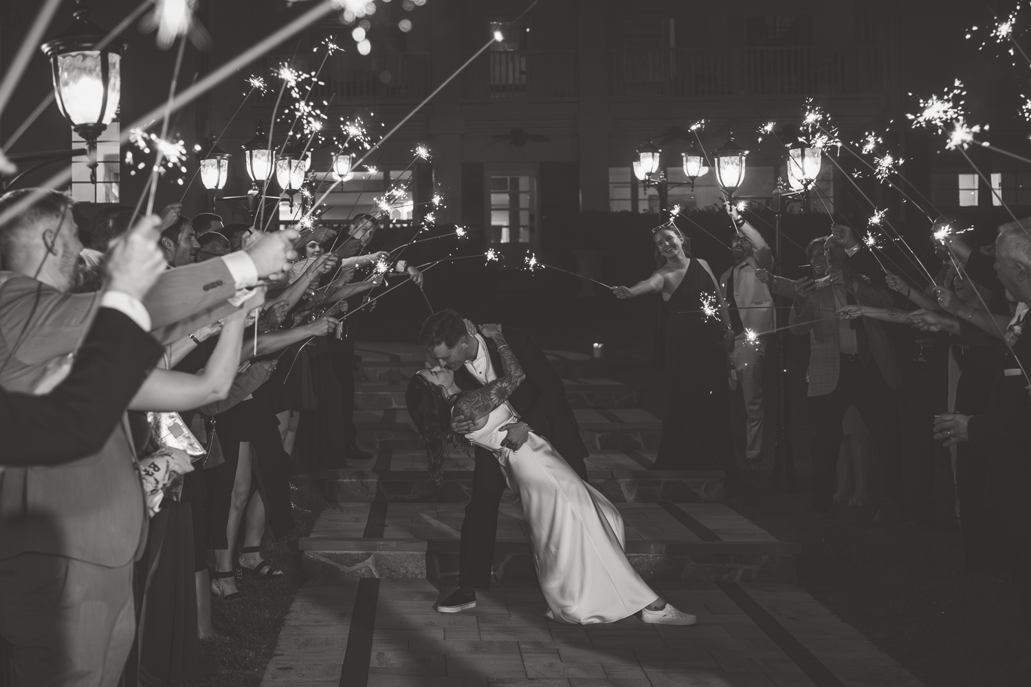 A couple shares a romantic dip kiss under sparklers held by wedding guests during their nighttime send-off celebration.