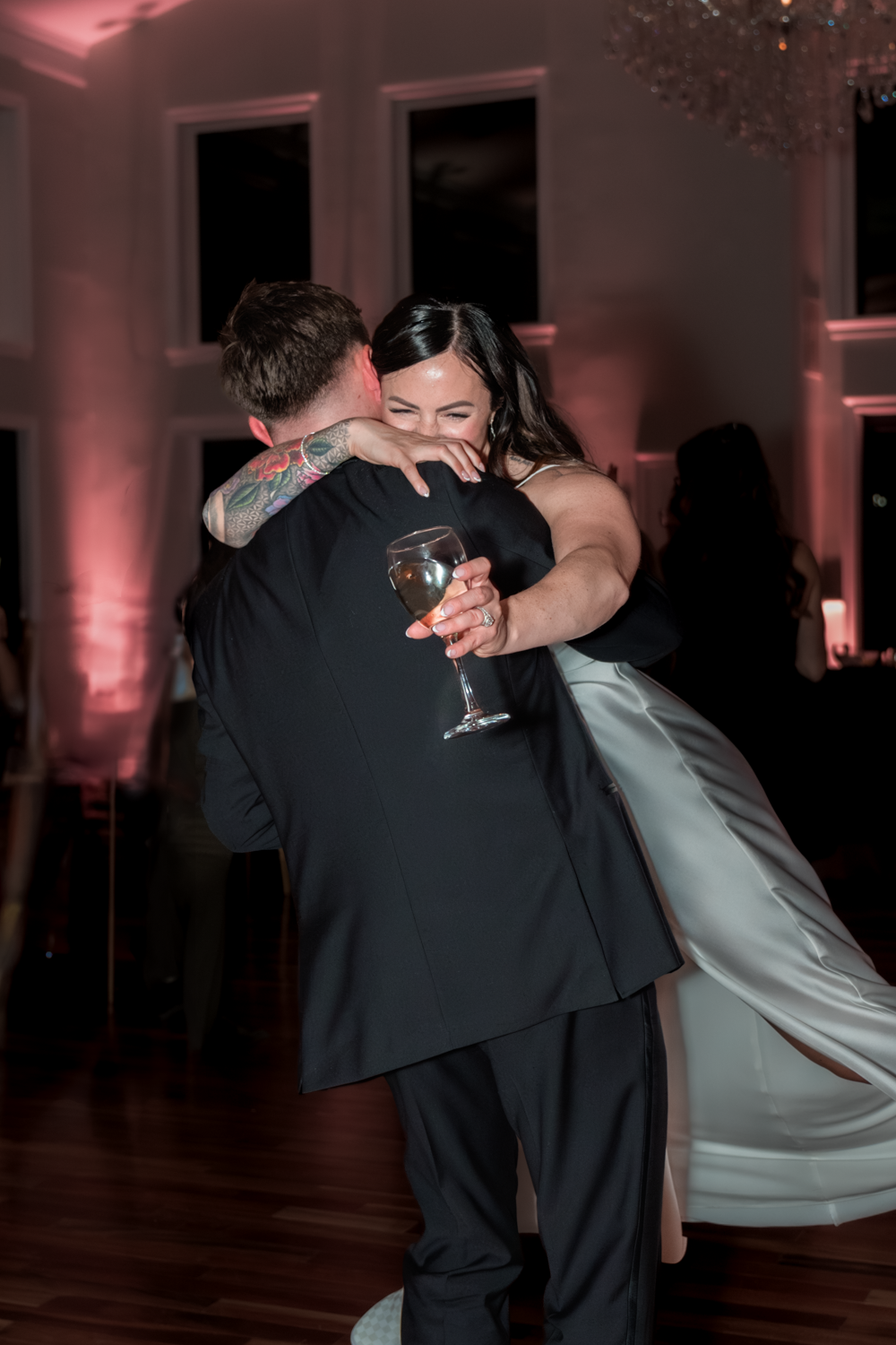 Couple in formal attire share a joyful embrace while dancing at an indoor reception with pink ambient lighting.