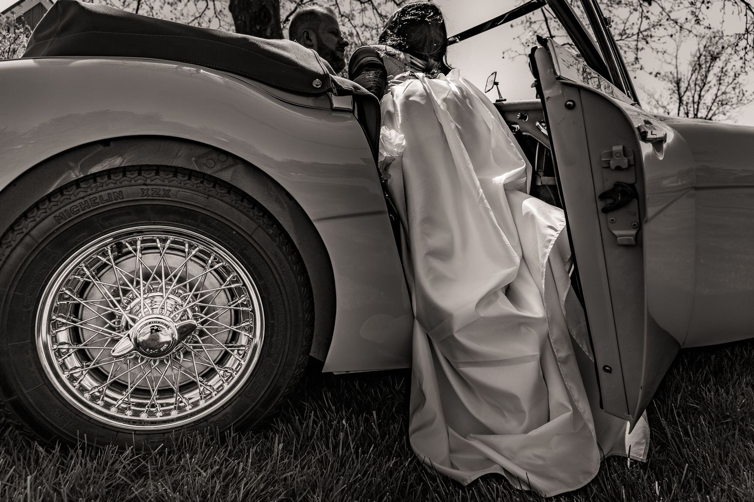 Black and white vintage photograph showing someone in flowing robes standing next to a classic car with wire wheels.