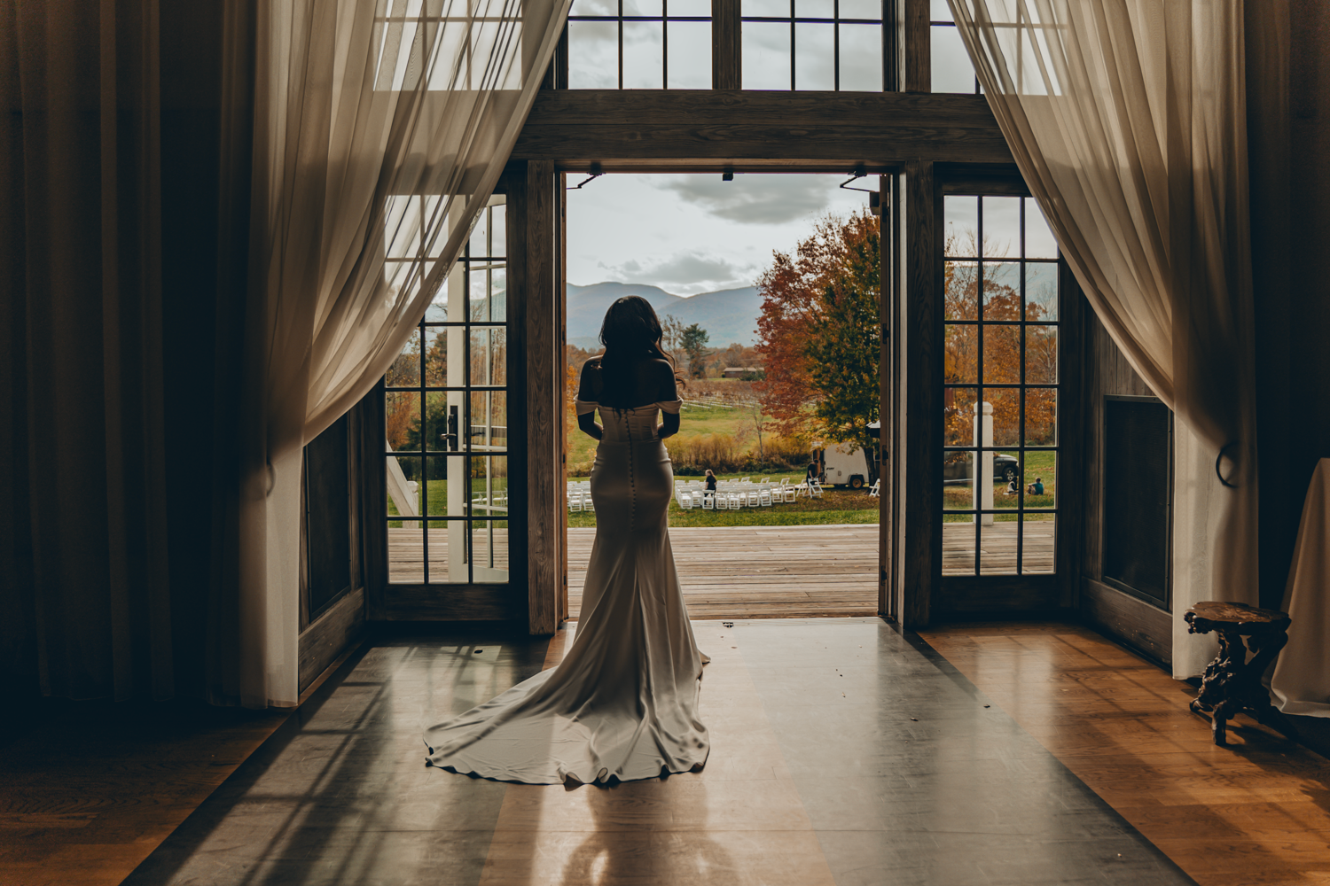 A bride in a white wedding dress stands silhouetted in elegant French doors overlooking an autumn landscape.