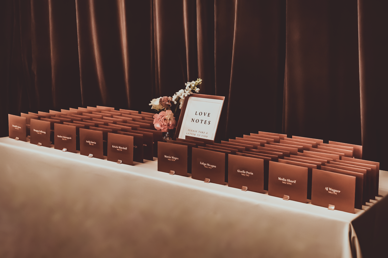 Rows of reddish-brown gift boxes are displayed on a white table with a small floral arrangement and sign.
