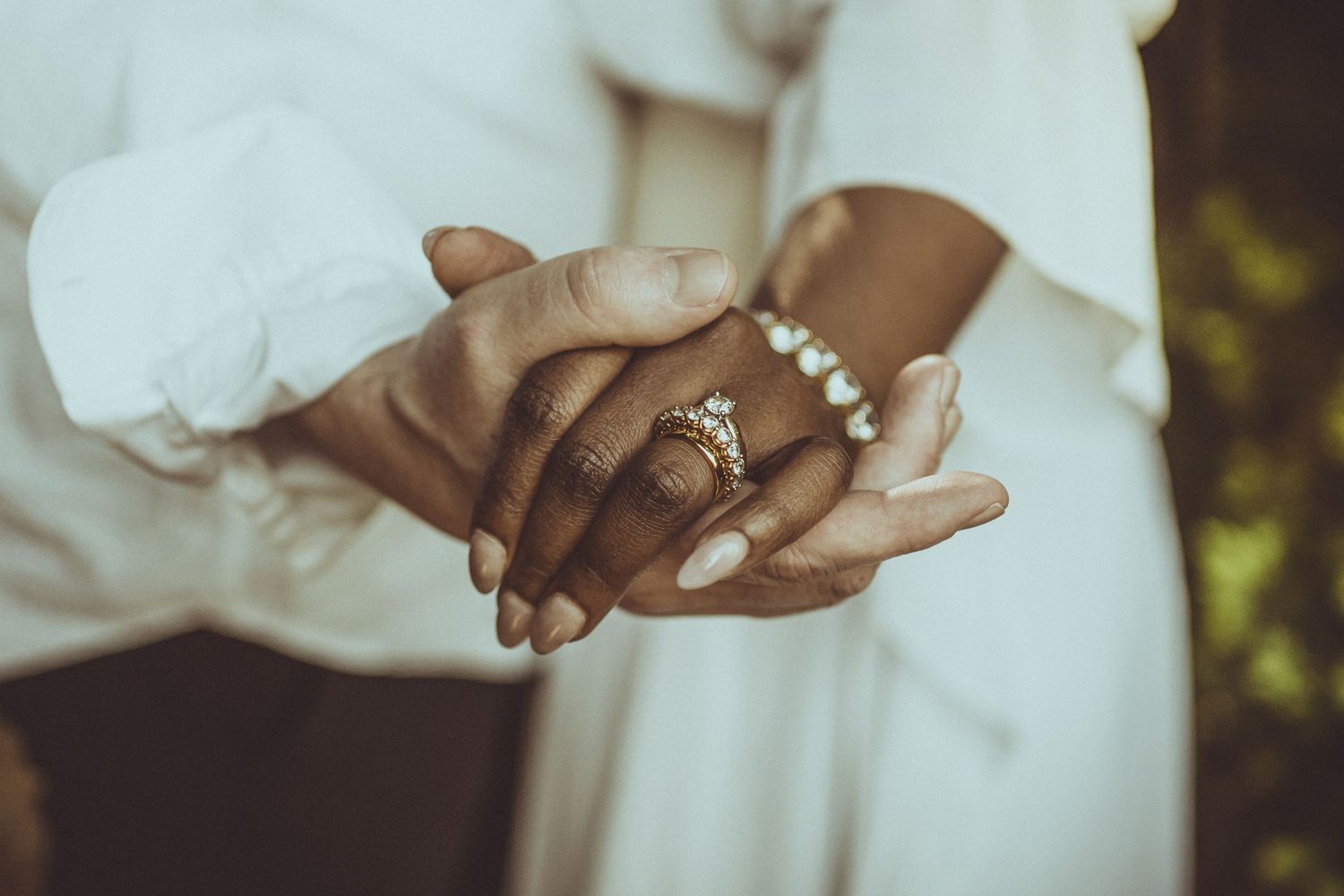 Close up detail of clasped hands wearing an engagement ring and delicate pearl bracelet against a white dress.