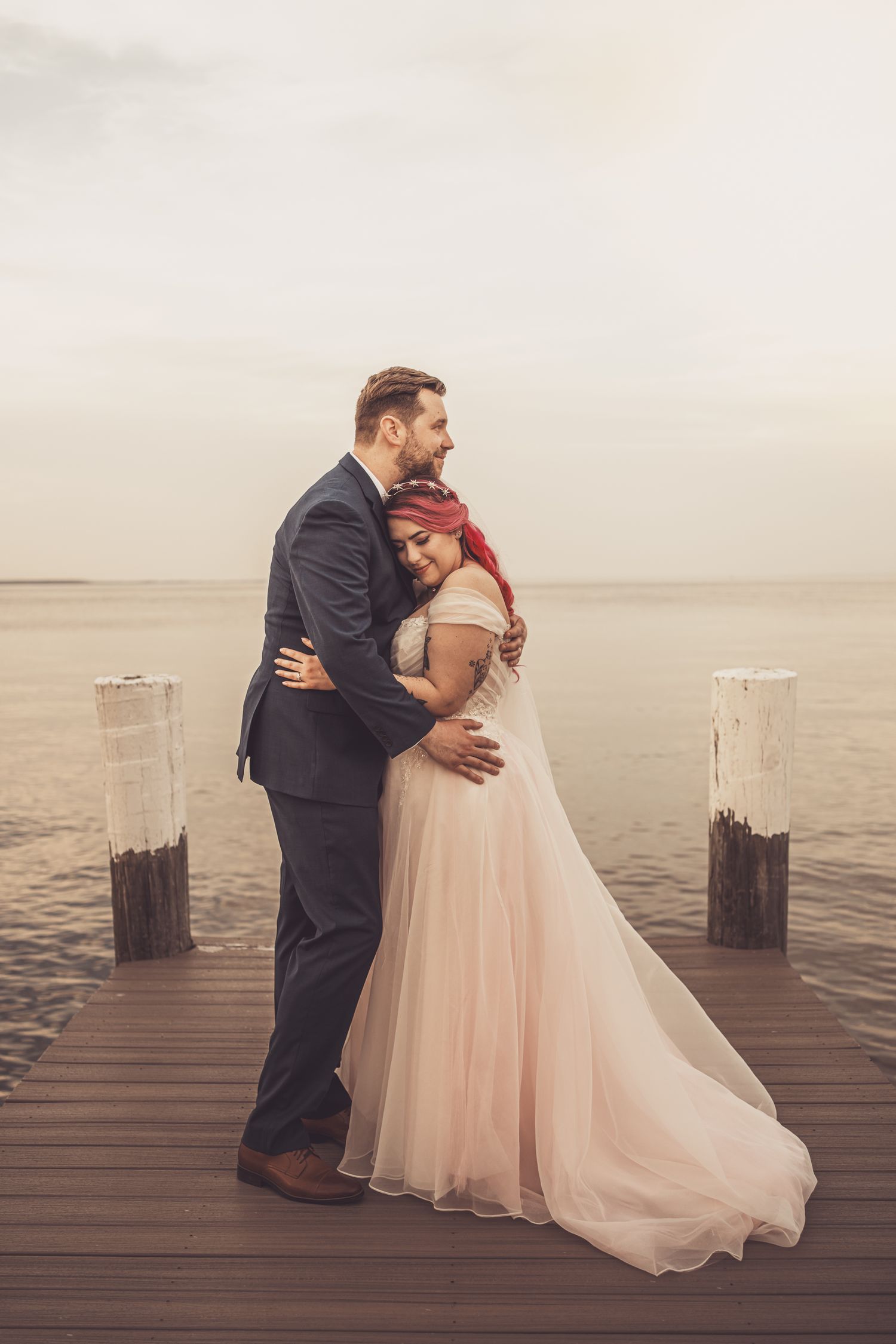 Romantic wedding couple embraces on wooden dock by water at sunset in ethereal peach gown and dark suit.