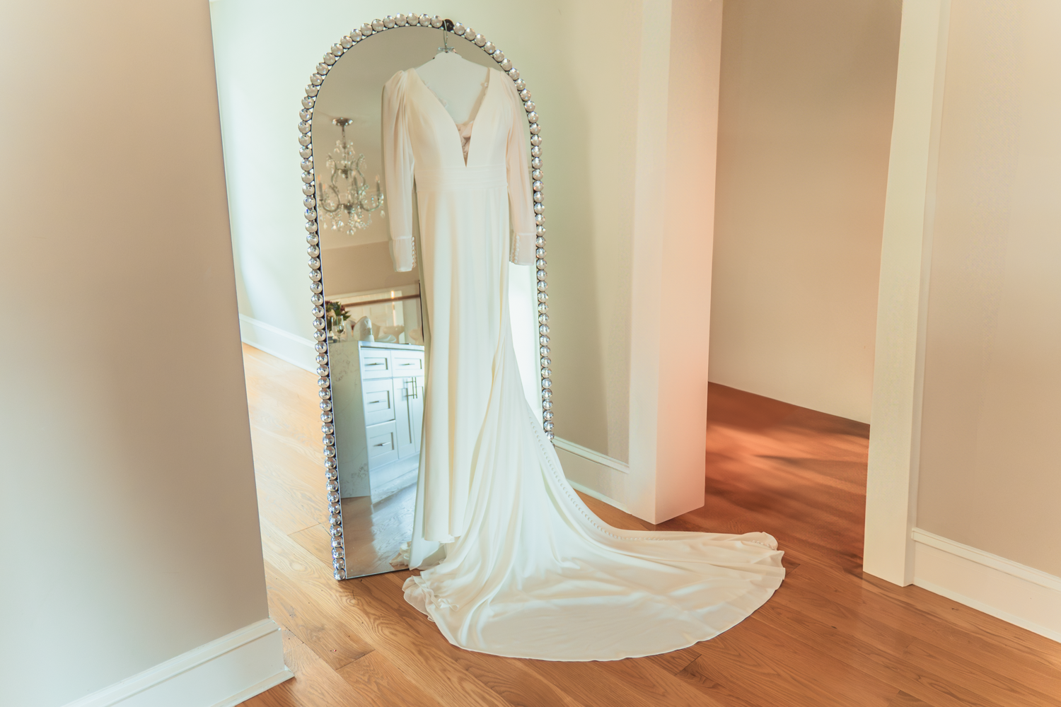 A flowing white wedding dress with a long train is displayed next to an ornate floor-length mirror on hardwood flooring.