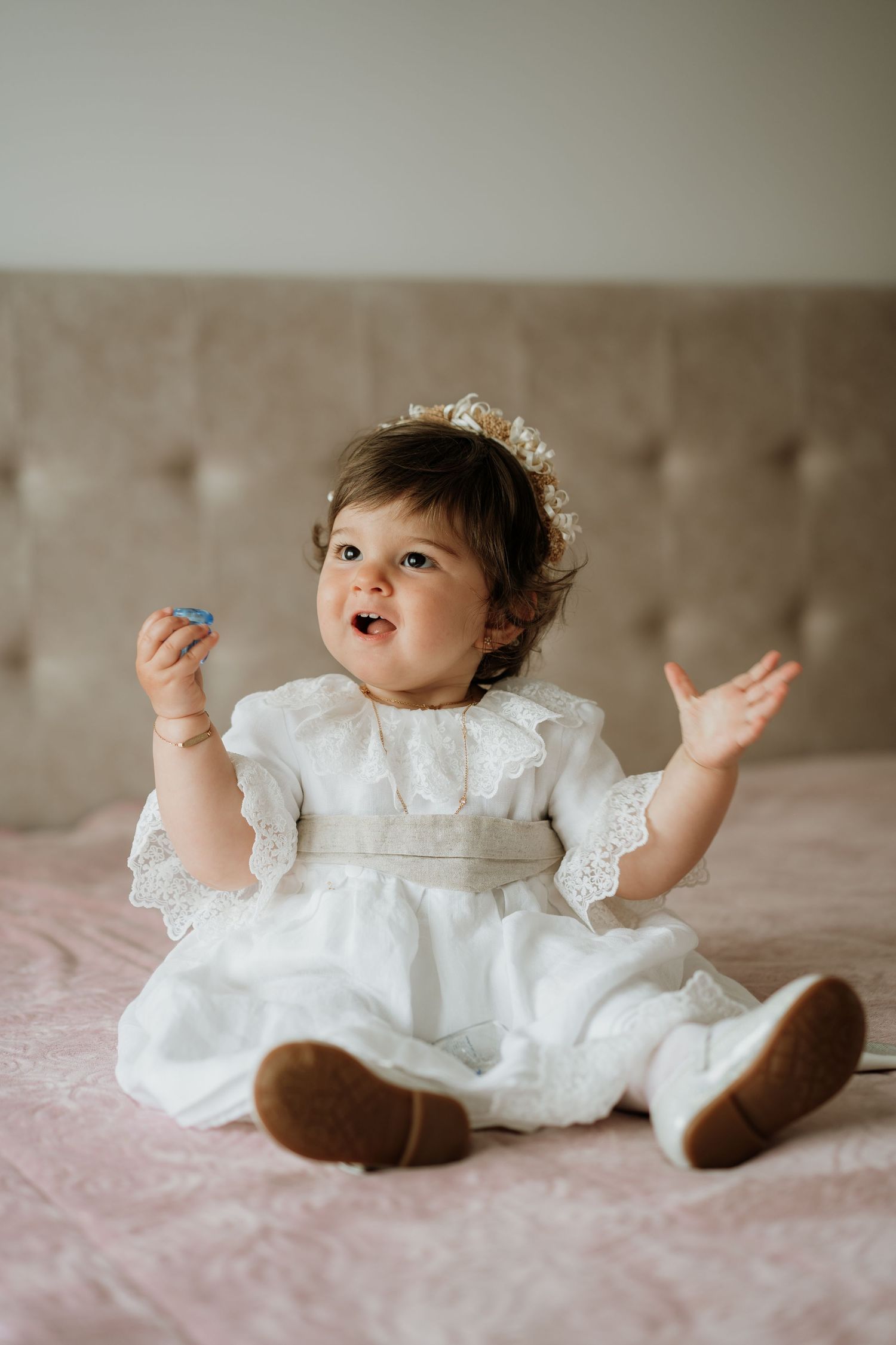 A baby girl in a white lace dress and floral headband sits on a pink bed, smiling with hands raised.