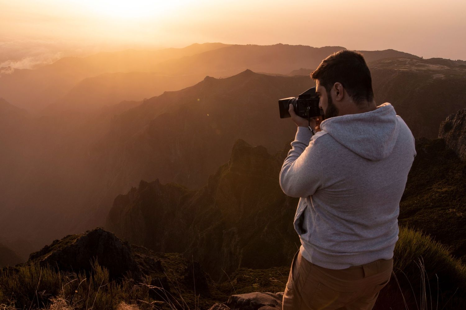 Photographer with camera captures stunning mountain landscape at golden sunset in hazy conditions.