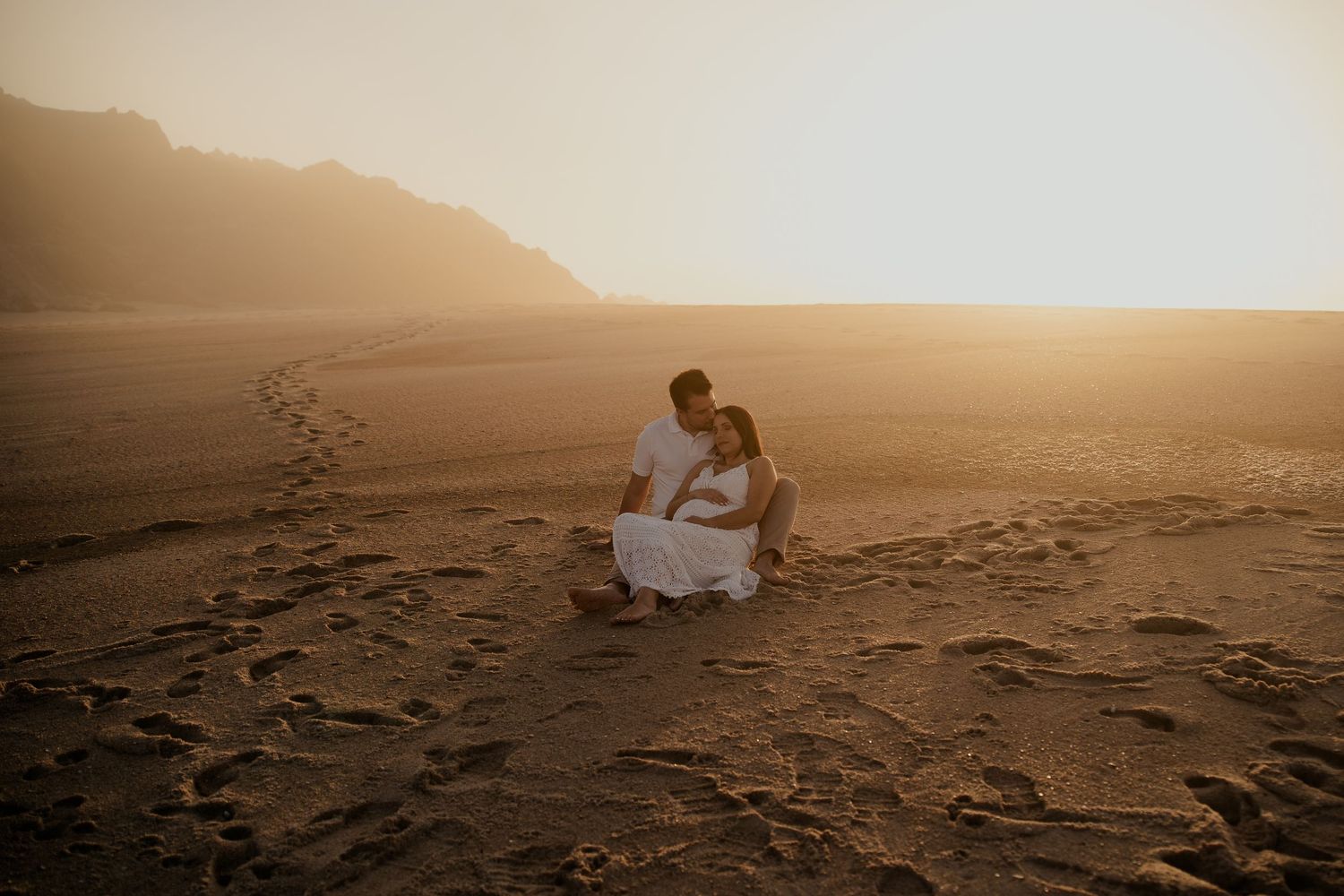 A couple shares a romantic moment at sunset on a sandy beach with mountains in the background.