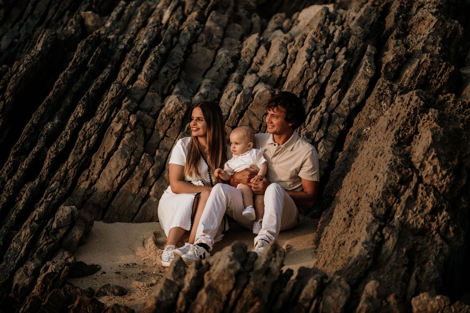 A happy young family with a baby sits against rocky cliffs on a sandy beach, all dressed in white outfits.