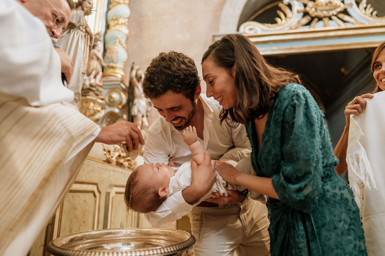 A priest baptizes a baby held by smiling parents at a church font during a christening ceremony.