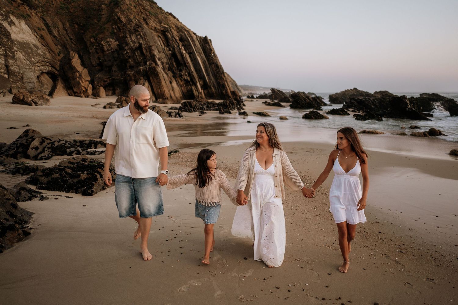 Family of four walking hand-in-hand on a scenic rocky beach at sunset, wearing casual white and denim outfits.