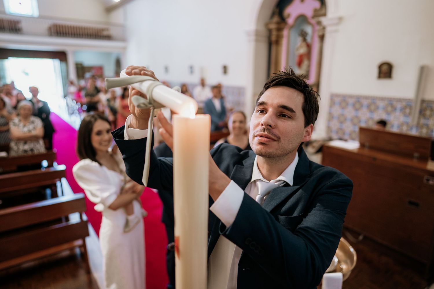 A groom in a navy suit lights a large white candle during a wedding ceremony inside a decorated church.