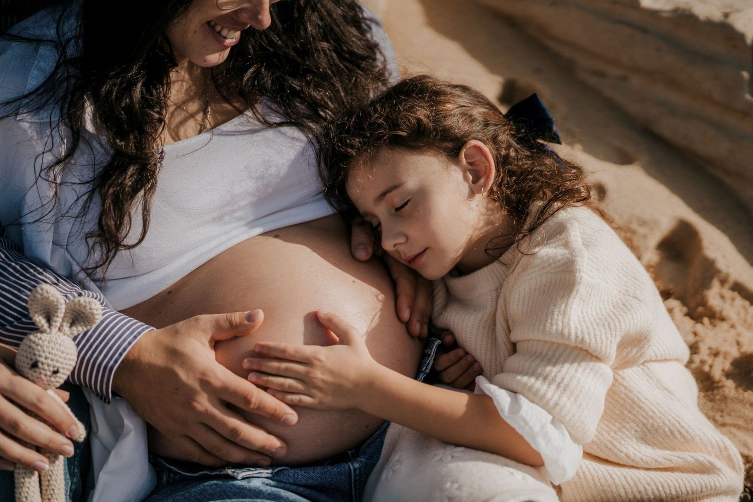 A young girl rests her head on her pregnant mother's belly while sitting together outdoors in a tender moment.