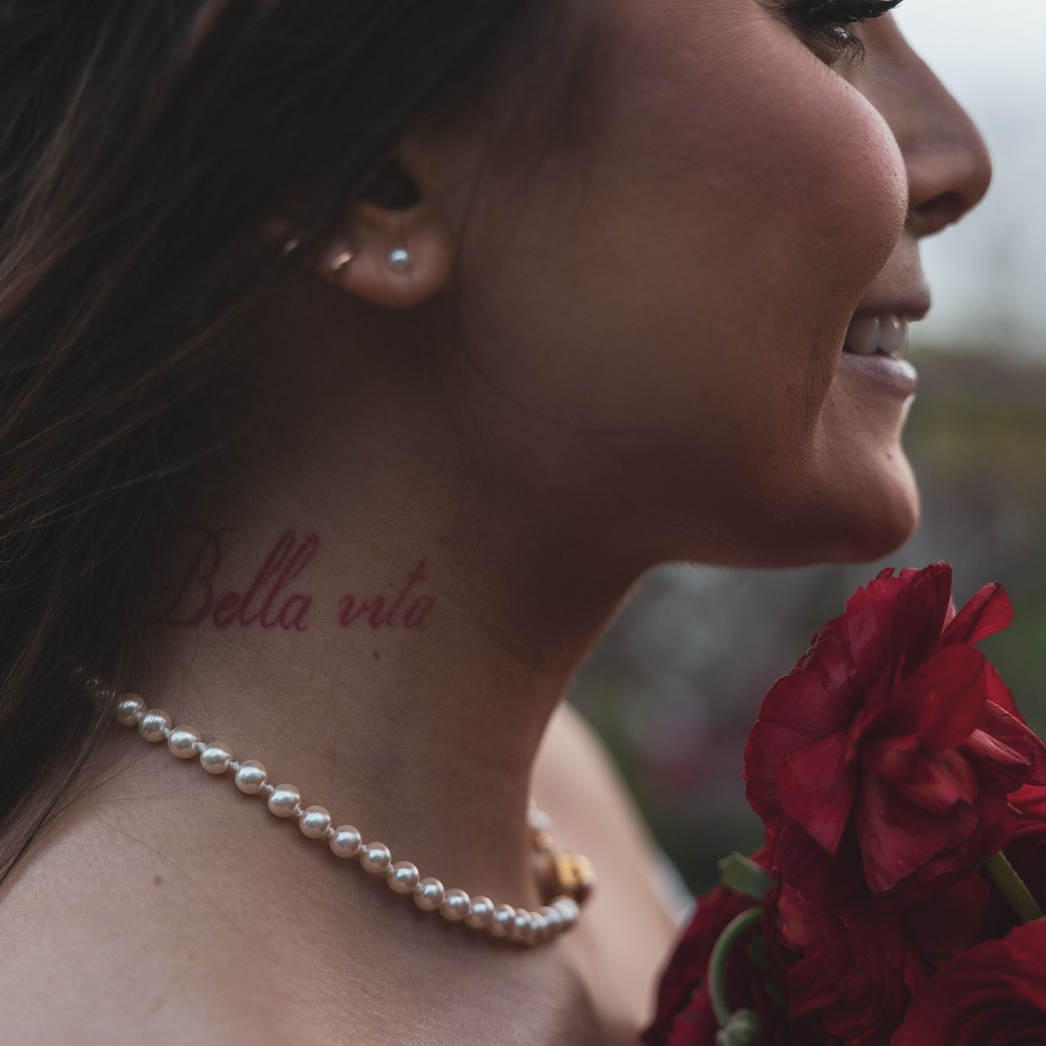 Close-up of person wearing pearl necklace with bella vita tattoo holding red rose flower.