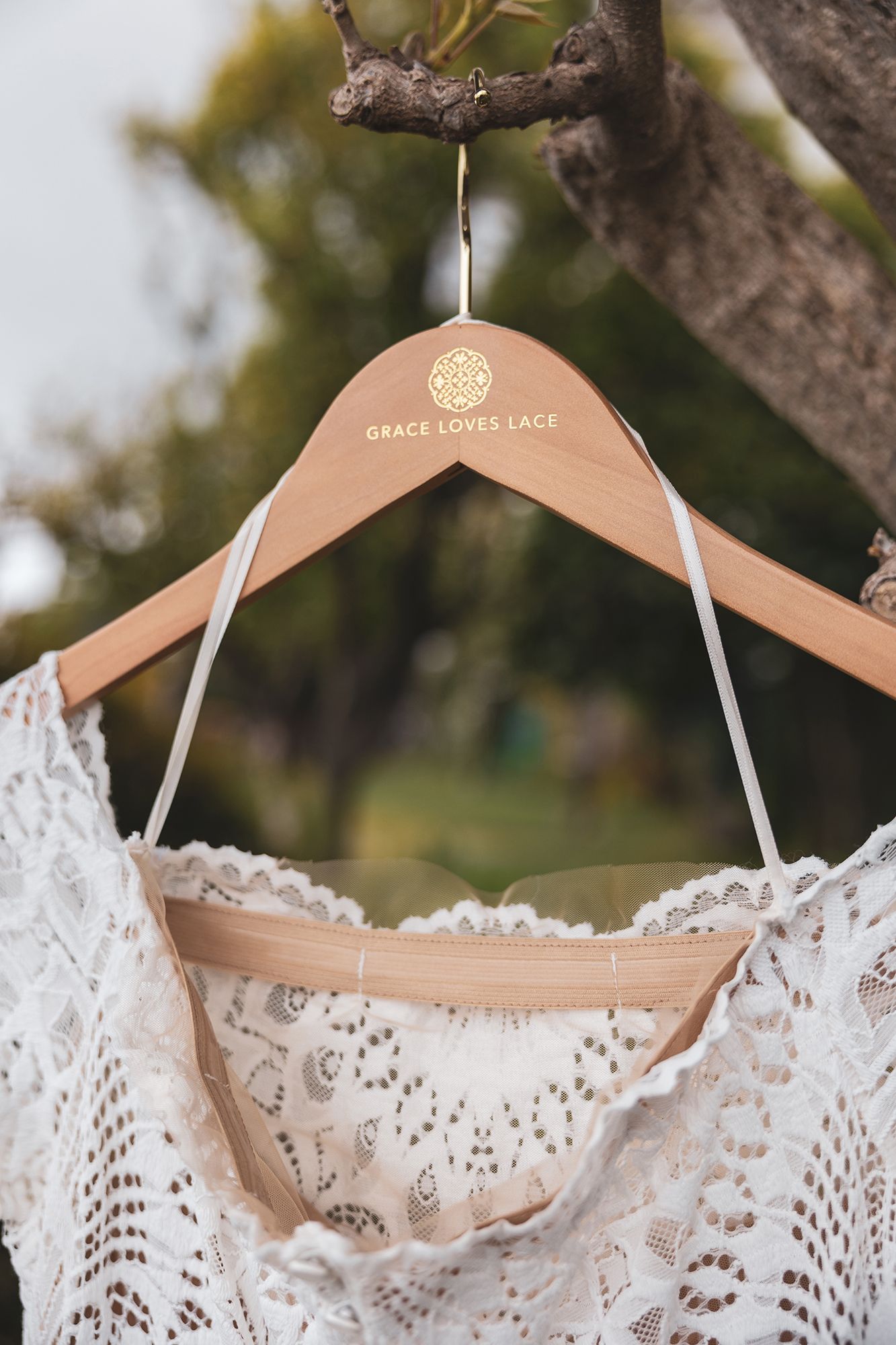 A delicate lace wedding dress hangs from a wooden hanger with 'Grace Loves Lace' branding against a blurred outdoor background.