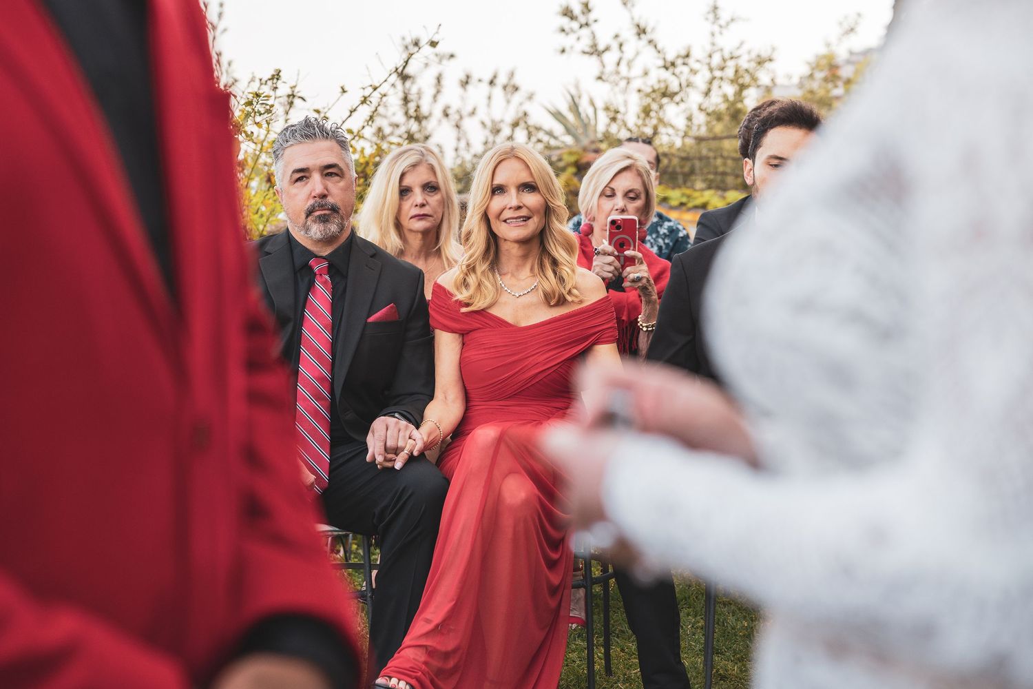 Wedding guests in formal attire gather outdoors, with a woman in a striking red dress as the focal point of the celebration.