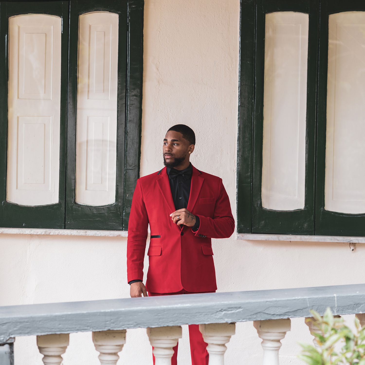 Person in a stylish red blazer and black turtleneck poses on a balcony with white pillars and green shutters.