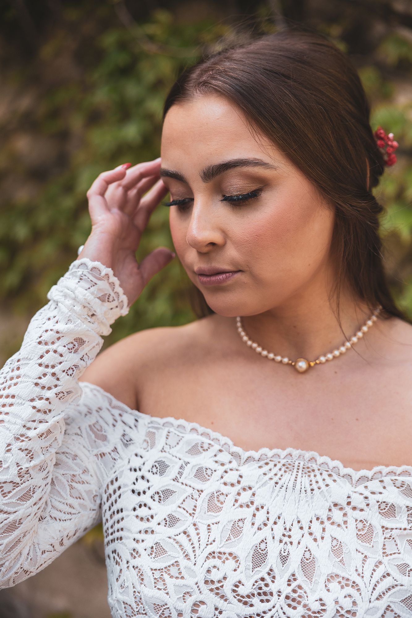 Portrait of someone wearing a white lace off-shoulder wedding dress with pearl necklace and delicate makeup in outdoor setting.