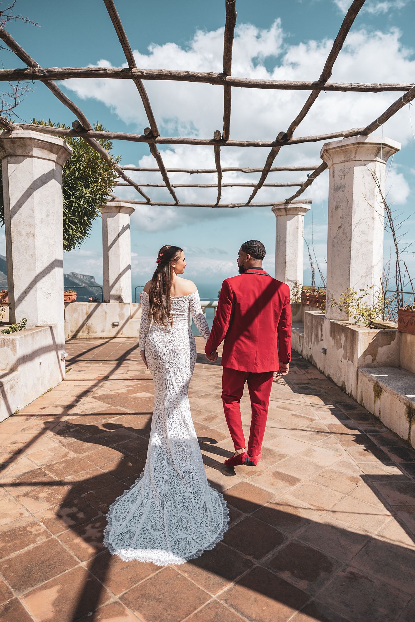 A couple in formal attire walking under a pergola overlooking a scenic coastal view at an Italian villa.