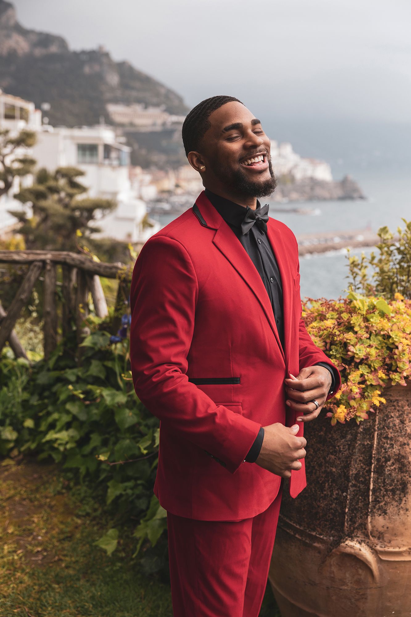 Person in a red suit smiles while standing on a scenic coastal terrace overlooking mountains and buildings in Italy.