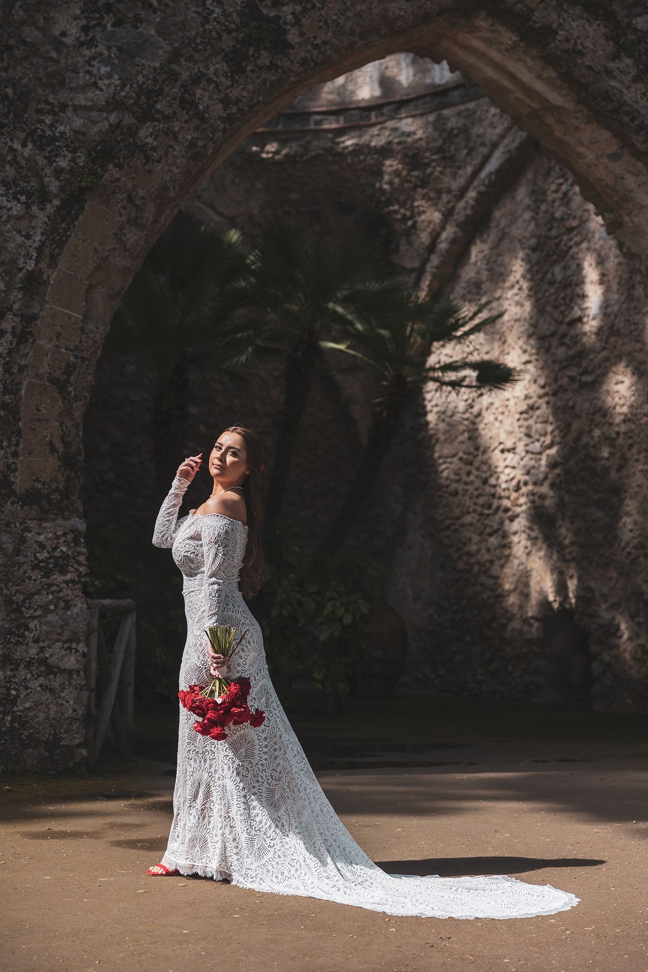 A figure in an elegant white lace wedding gown with a train poses beneath a stone archway in dramatic lighting.