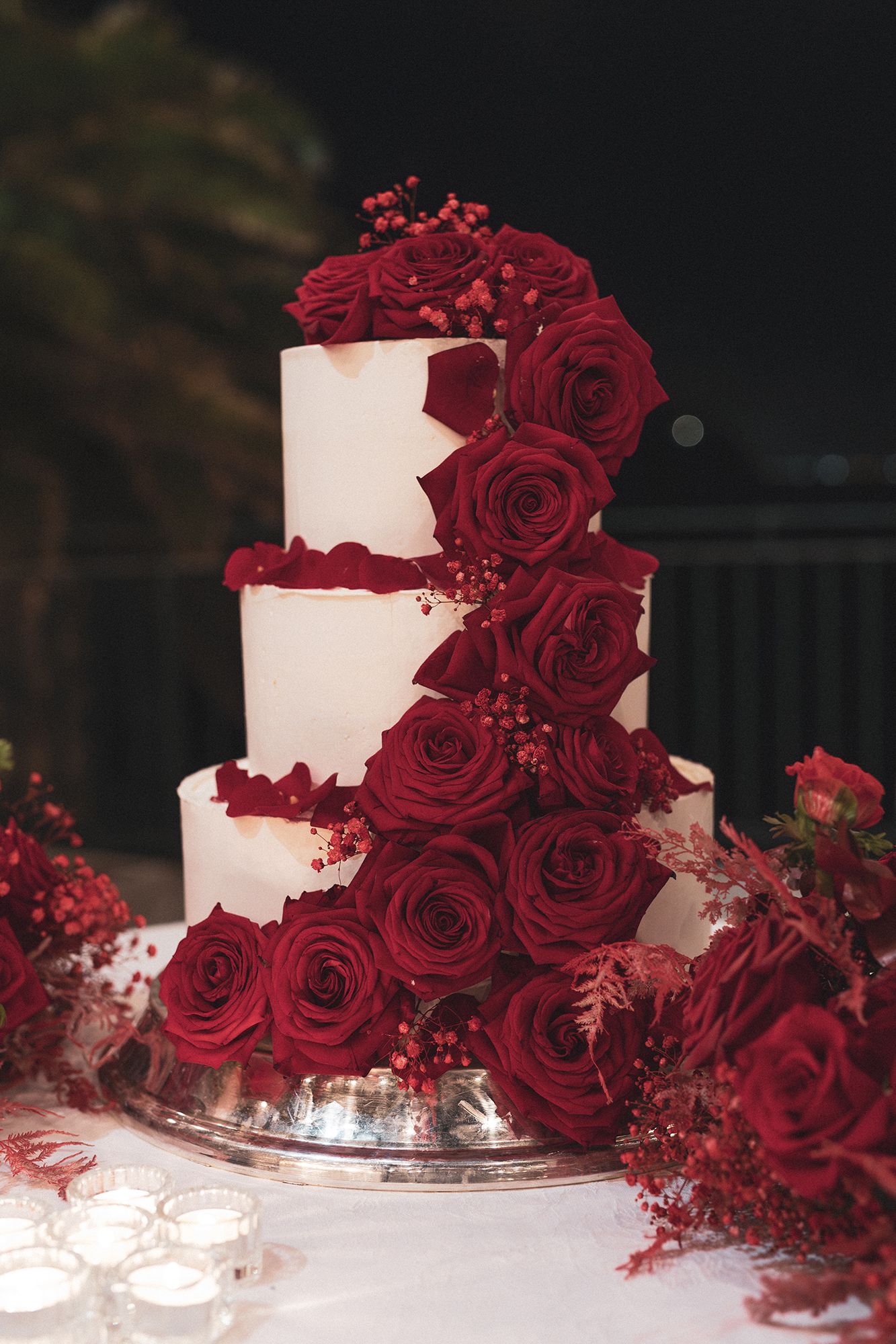 Elegant three-tier white wedding cake decorated with cascading deep red roses and floral accents on a silver stand.