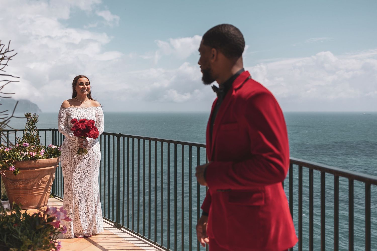 A couple shares a wedding first look moment on an oceanfront terrace with blue waters in the background.