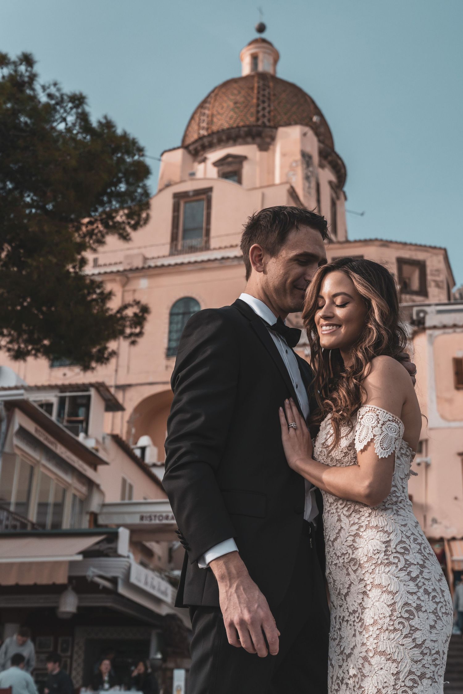 A couple shares romantic moments in formal attire outside a Spanish colonial style building with a domed roof at sunset.