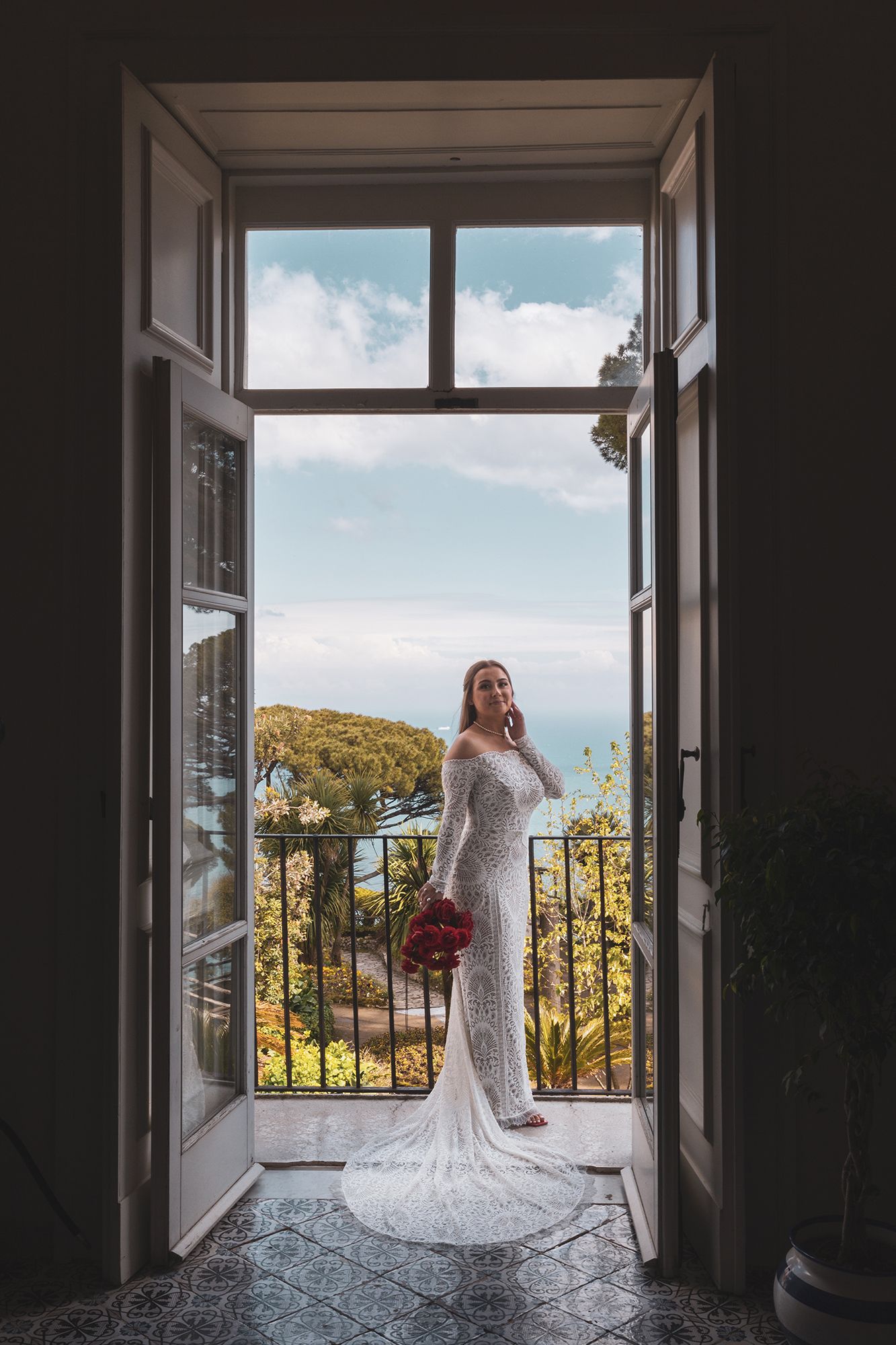 A bride in a lace wedding gown stands on a balcony overlooking the Mediterranean coast on a sunny day.