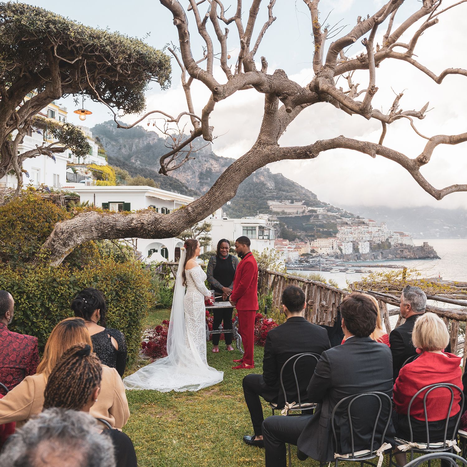 An outdoor wedding ceremony takes place under a twisted tree branch on a scenic cliff overlooking an Italian coastal town.