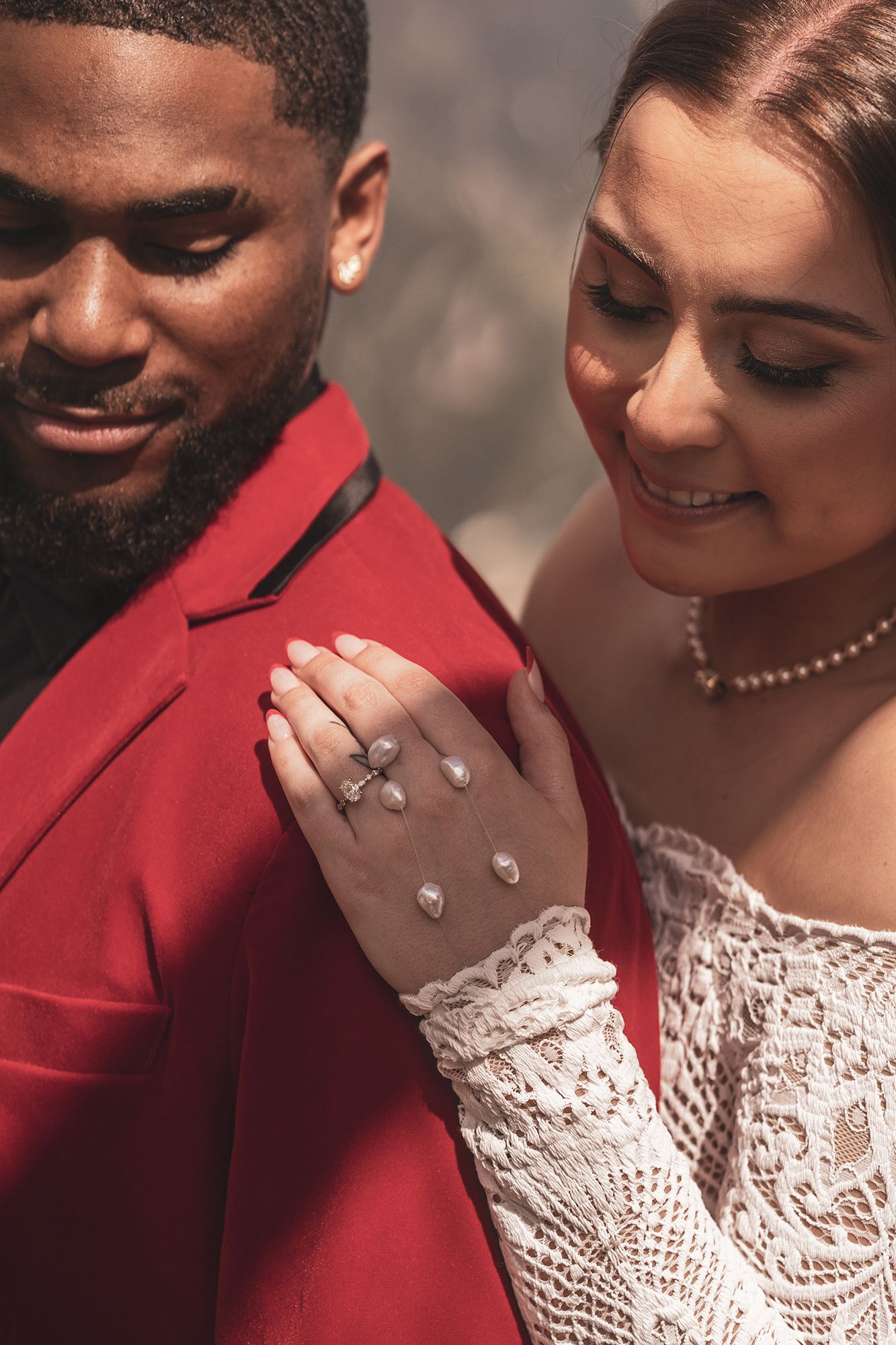 A romantic engagement photo with the couple smiling, showcasing a white lace dress, red suit jacket and sparkling ring.
