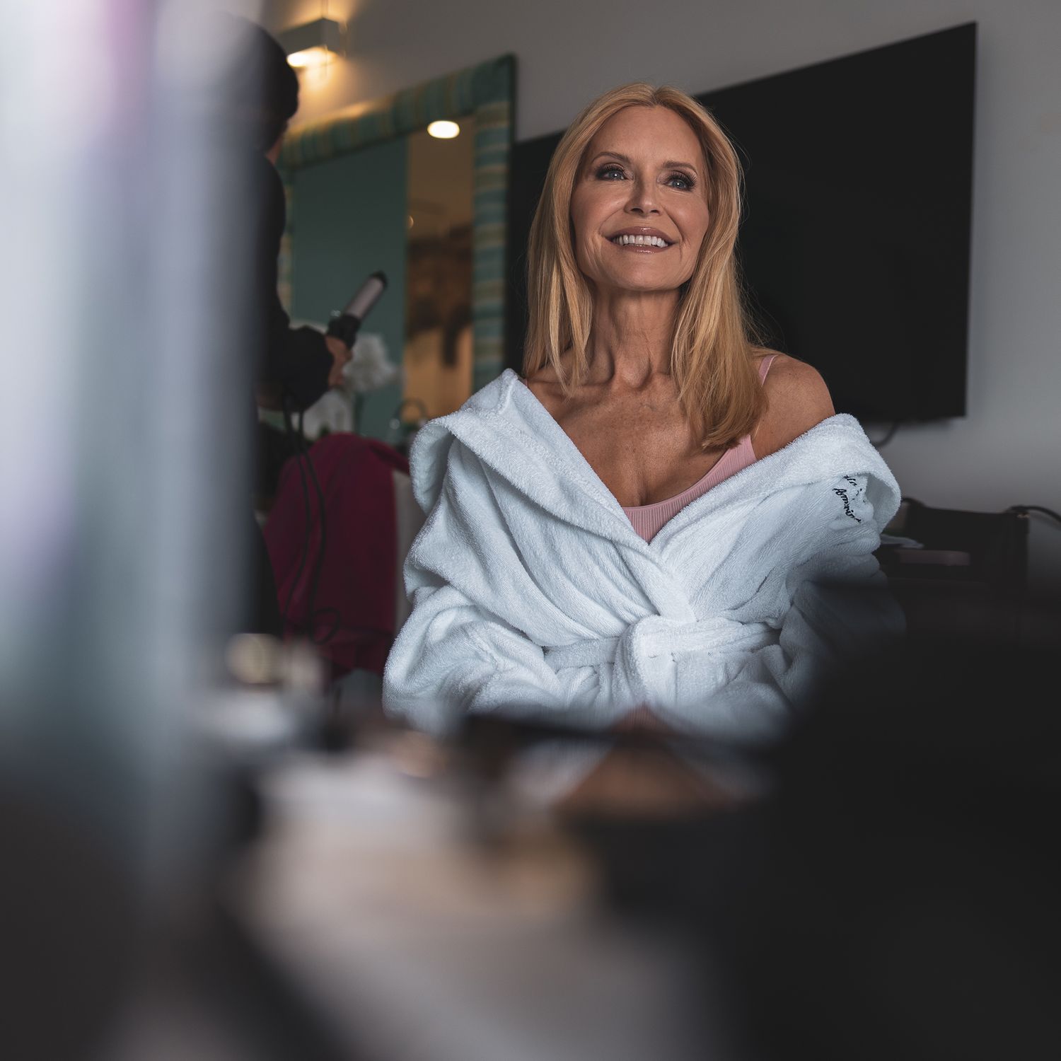 Person in a white bathrobe smiling while sitting in what appears to be a makeup chair.