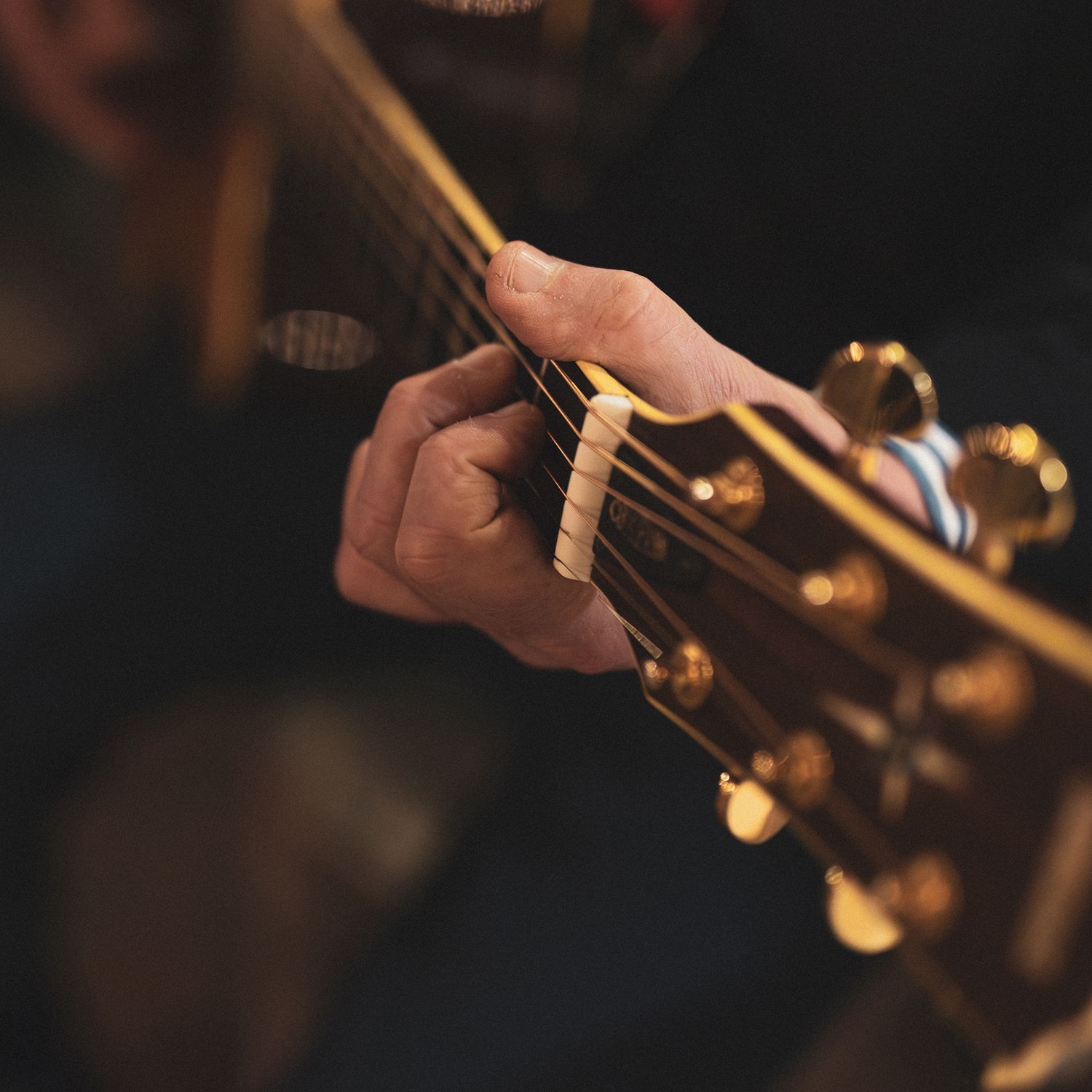 Close-up of hands playing acoustic guitar with golden tuning pegs visible in dark moody lighting.